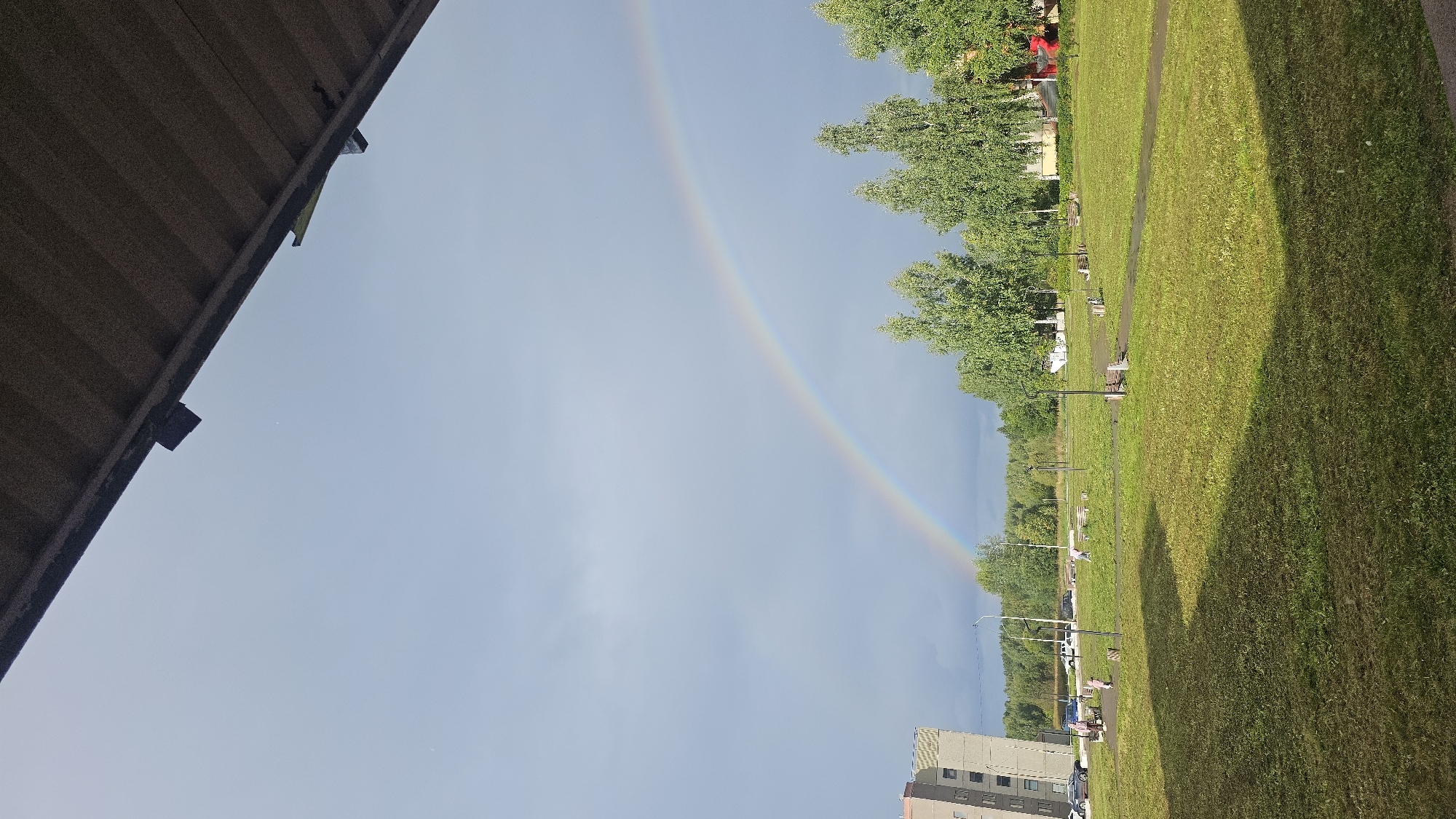 A double rainbow is visible in a cloudy sky over a green grassy area with scattered trees and buildings. In the foreground, a dark, corrugated roof overhang partially obscures the top of the image. To the left, a tall, multi-story apartment building is visible with several cars parked in front. The grassy area features a paved path, benches, and lampposts. Several people, appearing to be adults and children, are present in the grassy area, some walking or standing. The overall environment suggests a residential area within a city, likely in Марковский, Russia, given the architectural style of the building and the presence of birch trees. The lighting indicates it is daytime, with the sky suggesting recent rain.