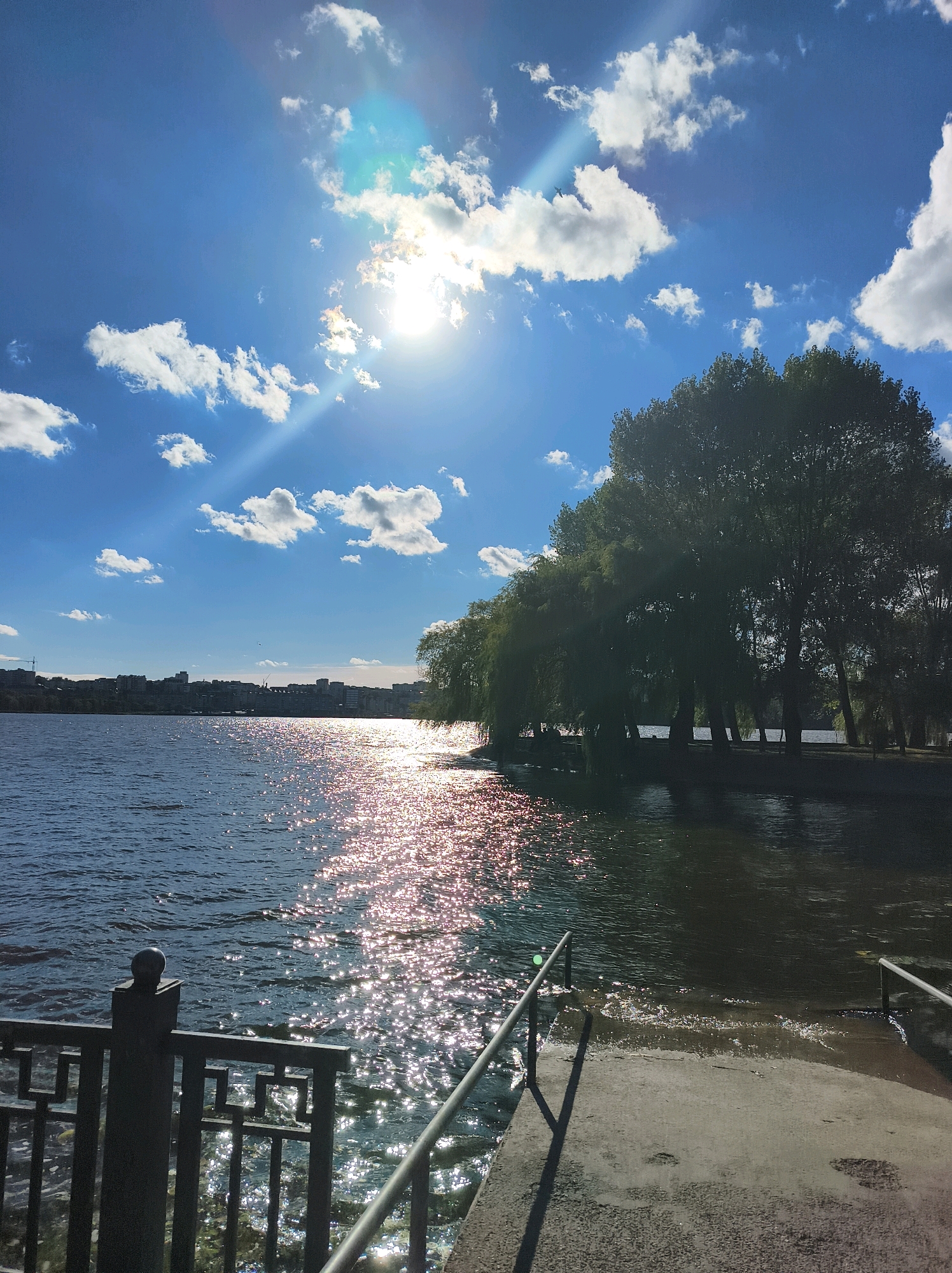 The image depicts a sunny day with a bright sun in a blue sky with scattered white clouds. The sun's rays create a glittering path on the surface of a body of water, likely Ternopil Lake in Ternopil, Ukraine. On the right side of the water, a park area with numerous tall trees is visible. In the background, a city skyline with buildings and a construction crane can be seen across the water. In the foreground, a metal railing and a concrete ramp leading into the water are present. The water appears calm with small ripples. The overall scene conveys a peaceful and bright atmosphere.