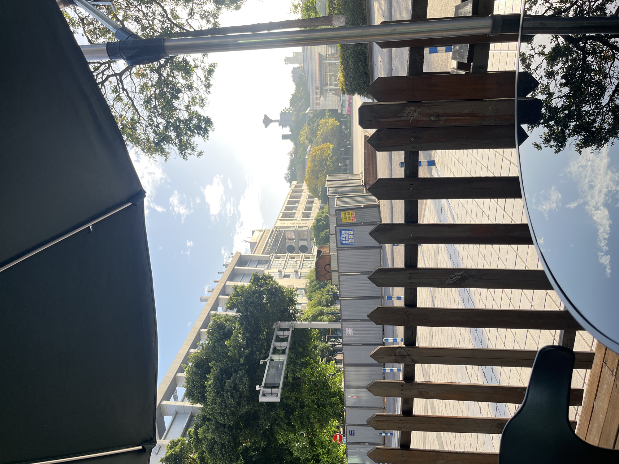 The image shows a view from under an umbrella, likely in an outdoor seating area. A wooden fence separates the seating area from a street with buildings in the background. Construction barriers are visible along the street. The table top of a table is reflecting the sky and clouds. The background buildings suggest an urban environment. Given the location, it's likely an area with commercial or residential buildings, possibly near a street undergoing construction or renovation. The scene gives the impression of a sunny day in Yuzhong District, China.