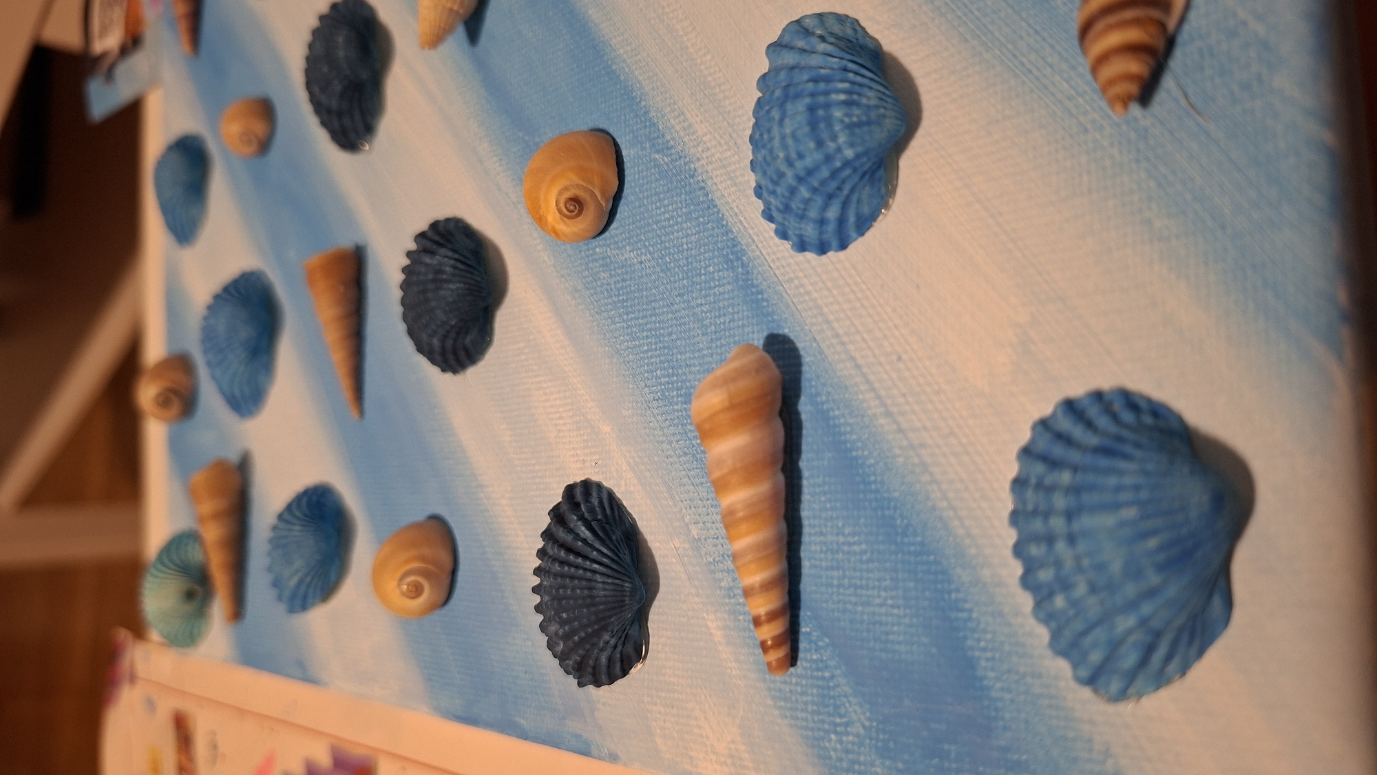 The image displays a canvas painted with diagonal light and dark blue stripes, mimicking waves. Scattered across the canvas are various seashells in different colors including blue, tan, and dark blue. Some shells are pointed, while others are rounded and fan-shaped. The scene is illuminated with a warm light, casting shadows from the shells onto the painted surface. The background is blurred but appears to be an indoor setting with furniture. There is no information to suggest this scene is located in Podgorica, Montenegro.