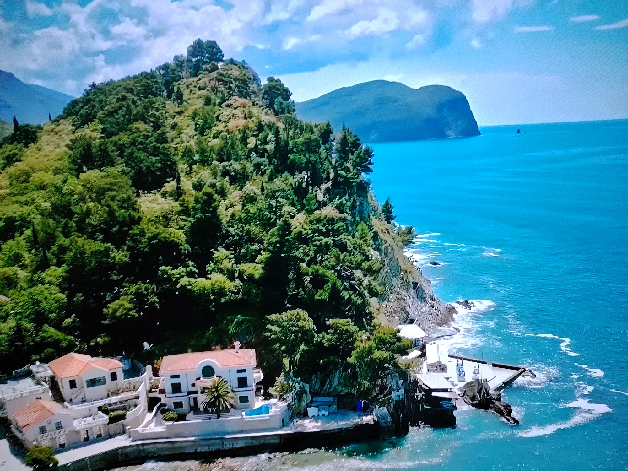 This is an aerial photograph of a coastal landscape. The scene is dominated by a verdant, tree-covered hill that slopes steeply down to the azure sea. Several white buildings with red-tiled roofs are situated at the base of the hill, near the water's edge. One building features a distinctive arched window and a palm tree in its courtyard. Adjacent to these structures, a modern-looking platform extends over the water. The sea is a vibrant blue, with white foam and waves lapping against the rocky coastline and the platform. In the distance, a large, dark, rounded hill or mountain rises from the sea. The sky above is a clear blue with scattered white clouds. The location is not in Huntingdonshire, United Kingdom; it appears to be a Mediterranean or similar coastal region.