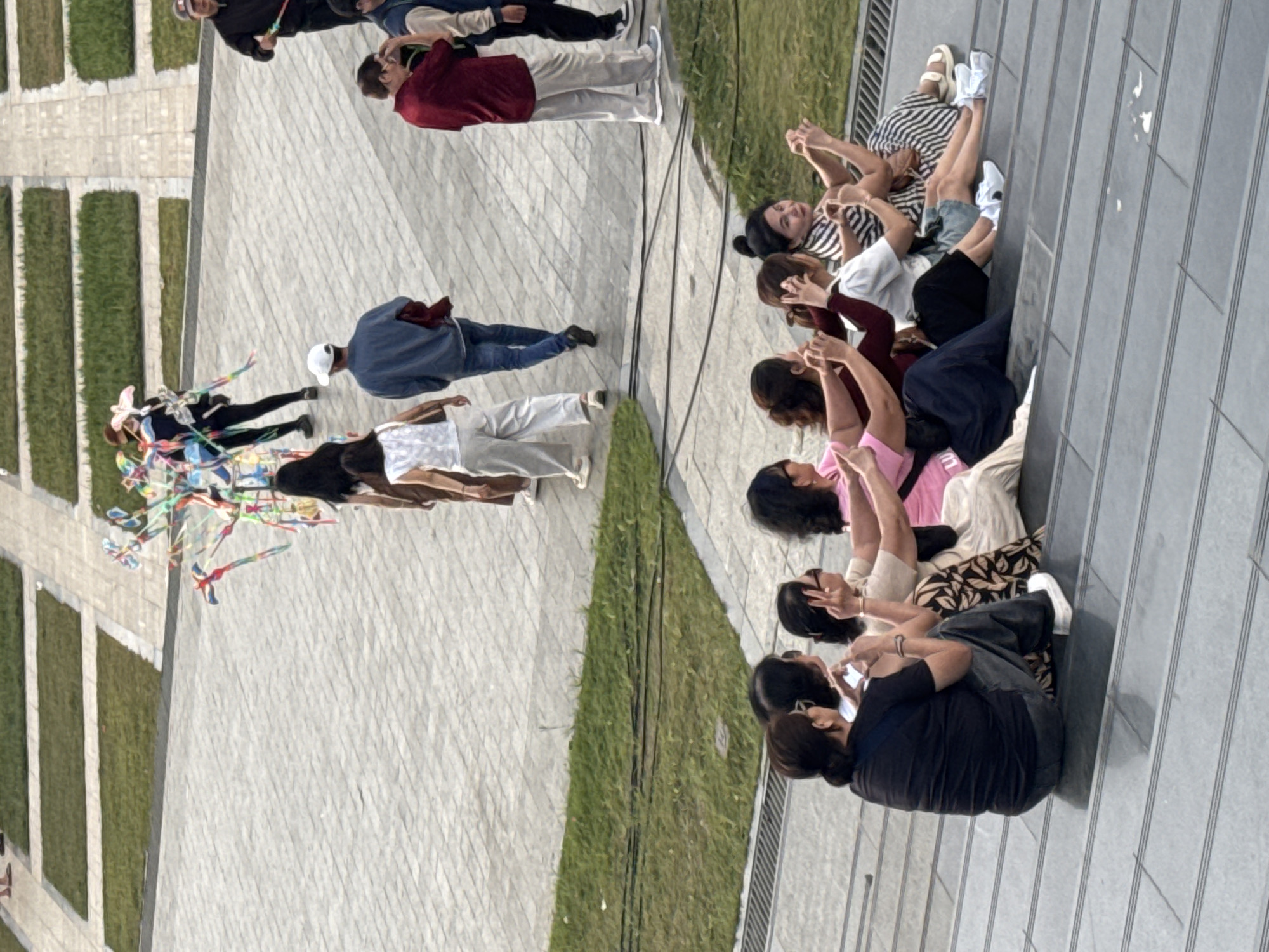 In Da Lat, Vietnam, a group of people are observed in a public outdoor space. In the foreground, seven women are seated in a row on tiered grey steps, facing away from the camera. Several of them are holding up their phones, appearing to take pictures or record videos. The women are dressed in casual clothing, including tops and skirts, with varying hairstyles and accessories. The background reveals a paved area with patches of green grass, suggesting a park or plaza. Further in the background, several individuals are walking on the paved surface. One person is holding a colorful kite, and another is carrying a red garment. The overall scene depicts a leisurely day in a public area, with people engaging in recreational activities and enjoying the outdoors.