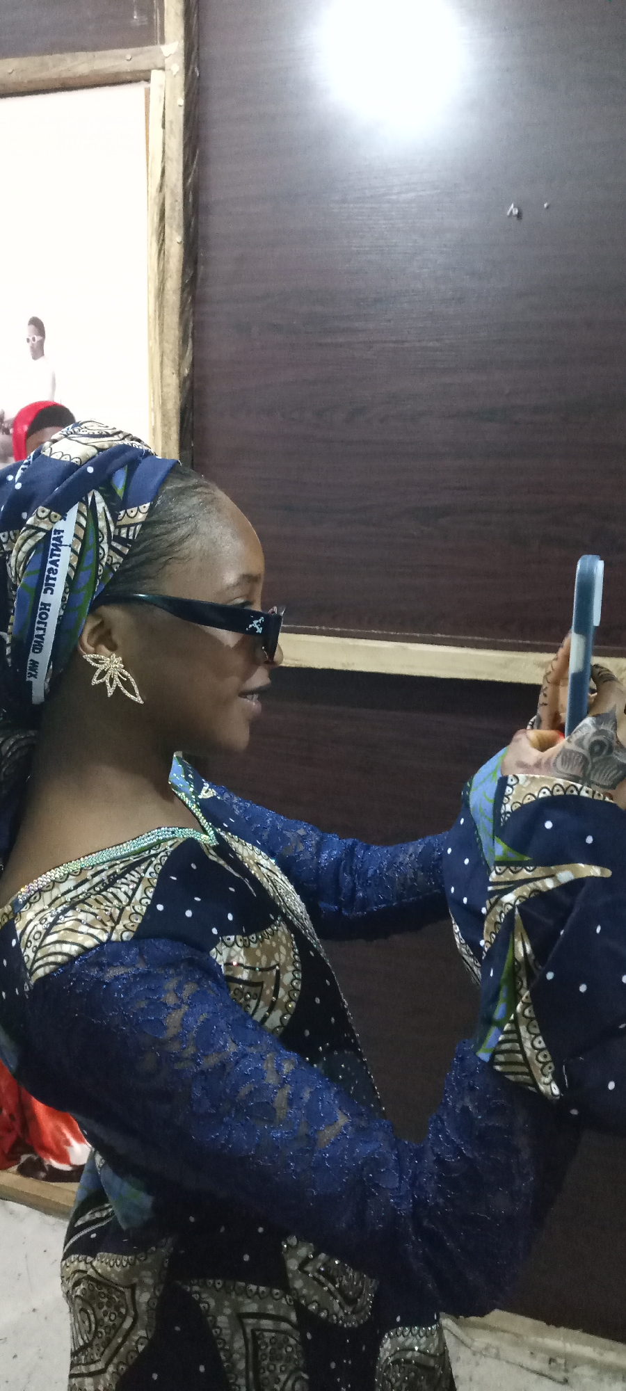 A woman wearing a blue and gold patterned dress, a matching headwrap, sunglasses, and leaf-shaped earrings is holding a smartphone in her right hand. The location appears to be indoors in an unknown city. There is a framed picture on the wall behind her, and a brown wall to her right.