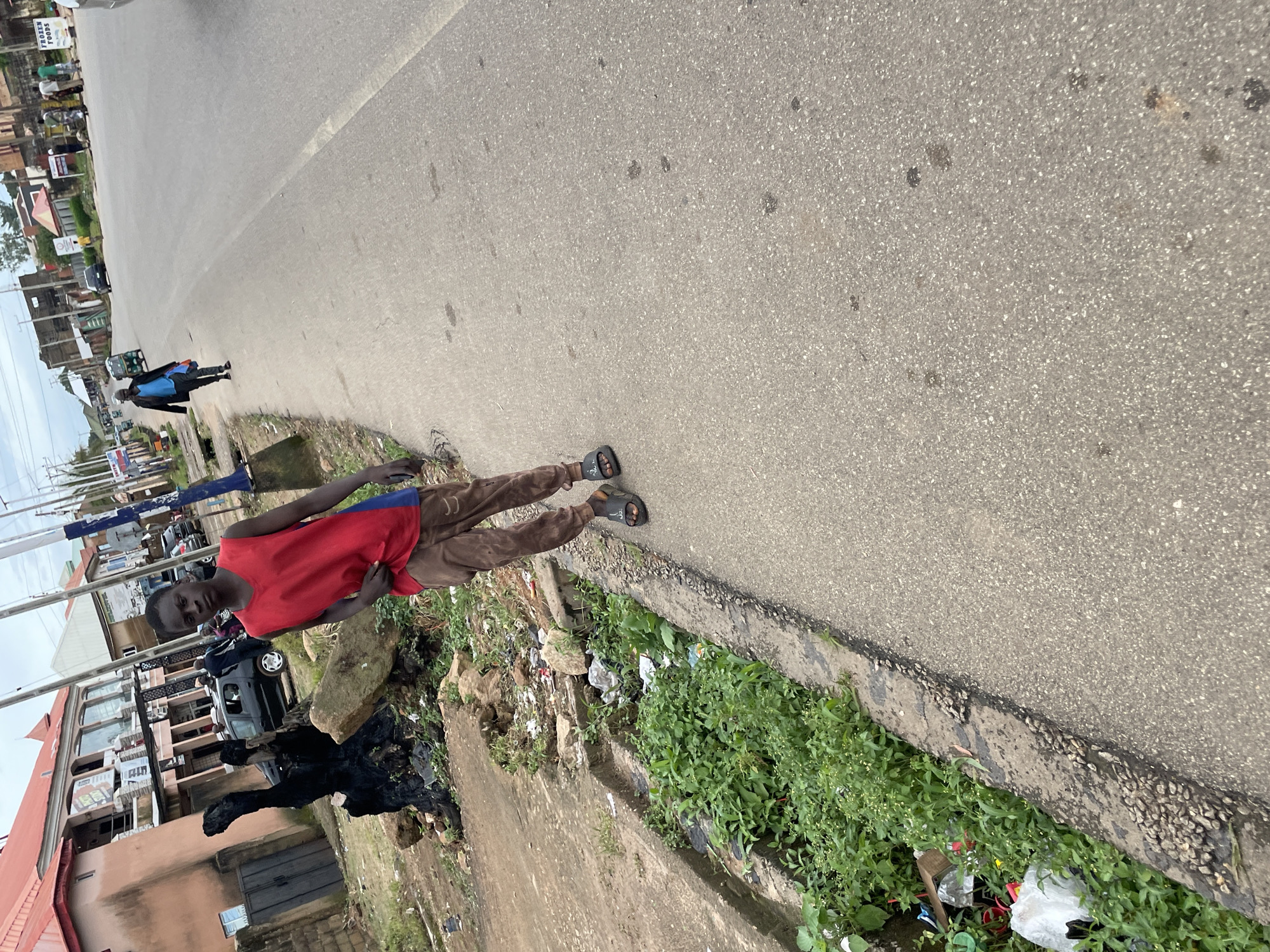 A young Black male stands on a paved road in Jos, Nigeria. He is wearing a red sleeveless shirt with a blue accent on the side, and brown sweatpants. He is also wearing black sandals. He is looking at the camera and has his right hand near his shirt. In the background, there are buildings and other people walking. The road is lined with a drainage ditch filled with green vegetation and trash. The area appears to be a mixed-use urban environment with commercial and residential buildings. The sky is overcast, suggesting it may be a cloudy day. The lighting in the image is somewhat dim.