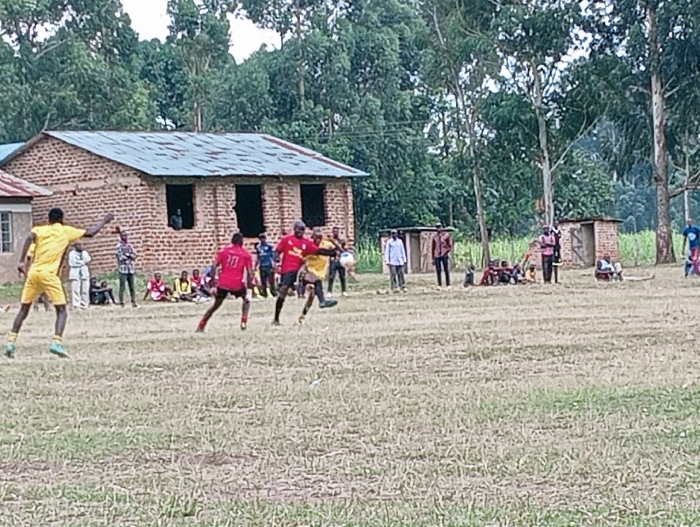 A soccer game unfolds on a dry, grassy field in a rural setting. Players in red and yellow jerseys compete, while spectators watch from the sidelines. A brick building with a tin roof serves as a backdrop, alongside lush trees and a small shed. The atmosphere suggests a local or amateur game, with a community gathering to enjoy the sport.