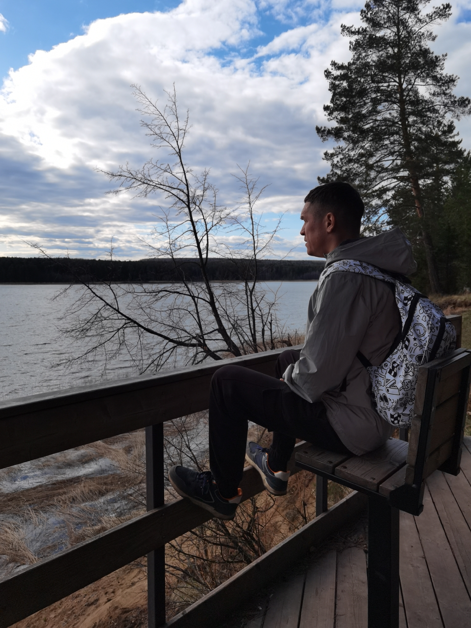 A man sits on a wooden bench on a wooden viewing platform overlooking a lake. The man has short hair and is wearing a grey hooded jacket, dark pants, and blue athletic shoes with orange accents. He is wearing a white backpack with cartoon-like illustrations. The lake is surrounded by trees, and the sky is cloudy. A leafless tree branch is in the foreground. The location is most likely in Votkinsk, Russia based on the request.