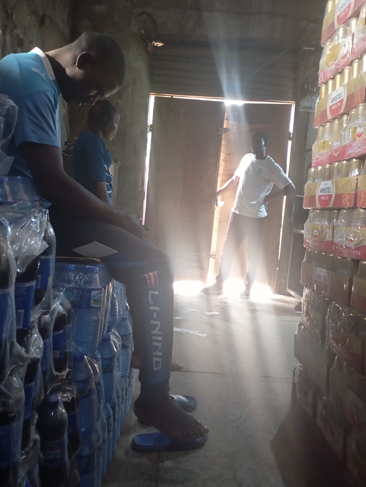 In a dimly lit storage facility, likely a warehouse in Shendam, Nigeria, a man in a blue t-shirt is seated on stacked cases of bottled beverages. Beside him, another person in a blue shirt is visible. The stacked cases are wrapped in plastic and appear to be water bottles, with some darker bottles, possibly soda, at the bottom. In the background, a doorway is open, allowing shafts of sunlight to illuminate dust particles in the air. A second man, wearing a white t-shirt and dark pants, stands near the doorway, with his hands on his hips, looking towards the interior of the facility. To the right of the frame, tall stacks of canned beverages, identified by "AMSTEL MALTA" labels, are also shrink-wrapped and neatly arranged. The floor is concrete, with some scattered papers. The overall impression is a scene of commercial storage and distribution.