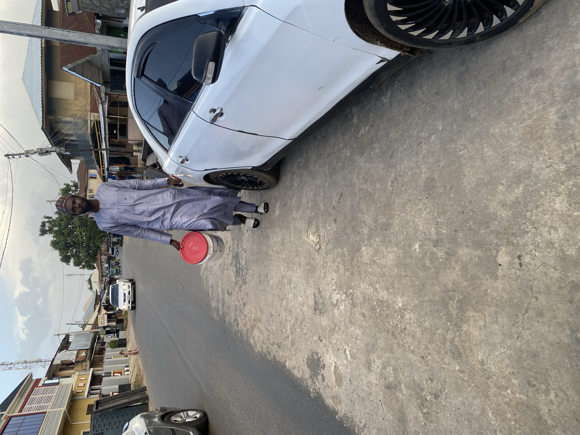 A man walks on a dusty, paved street in Jos, Nigeria, carrying a red-lidded white bucket in his left hand. He is wearing a long, light gray traditional outfit and a cap. A white sedan is parked to his right, and a silver car is visible in the background on the left. Buildings line the street, some appearing to be shops or homes. The sky is overcast. The scene appears to be a typical street view in a Nigerian city.