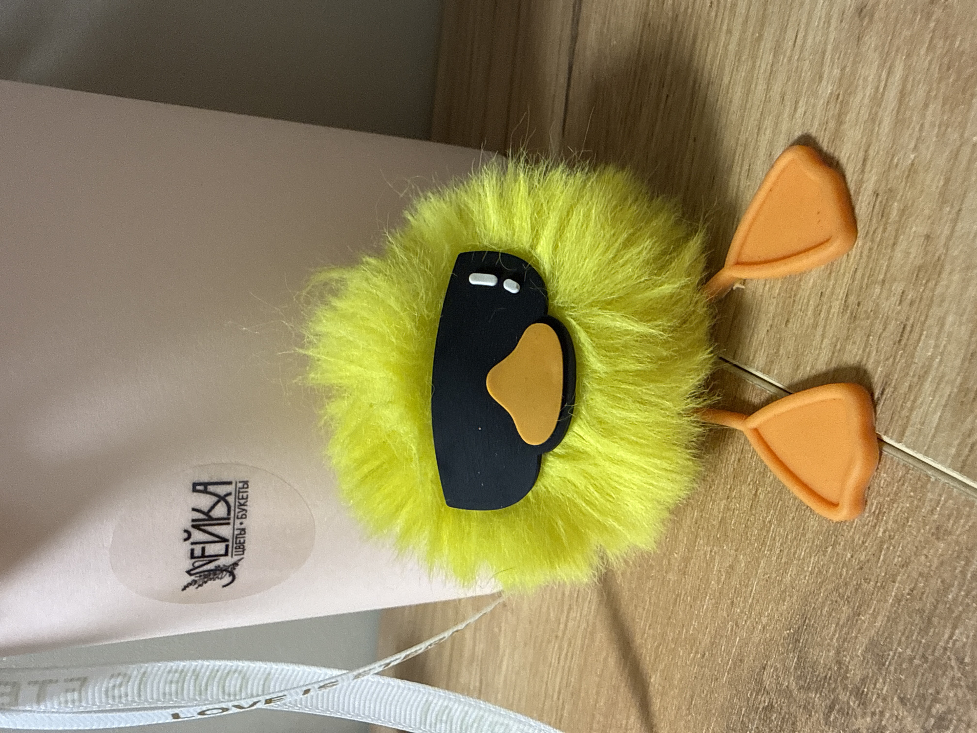 The image shows a yellow, fluffy chick decoration with orange feet and a black and orange beak, lying on a light brown, textured surface. A white gift box with a sticker in Russian "НЕЙКА цветы - букеты" is partially visible on the left. A white ribbon with the word "LOVE" is also partially visible. The photograph appears to have been taken indoors in Votkinsk, Russia, based on the company name in the sticker on the box.