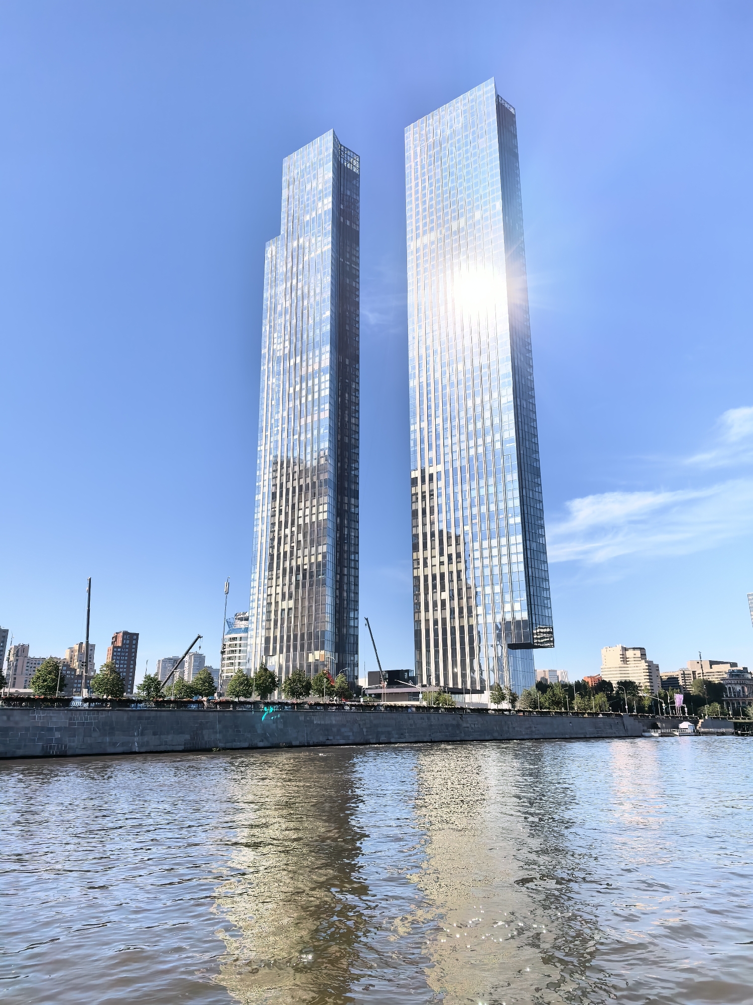 The image captures a bright, sunny day with a clear blue sky over the Moscow River. Two prominent, modern skyscrapers with glass facades dominate the scene, reflecting the sunlight and the surrounding cityscape. The river in the foreground has a rippling surface with reflections of the buildings and sky. Along the riverbank, a stone embankment is visible, lined with trees and other buildings. Construction cranes can be seen behind the skyscrapers, suggesting ongoing development in the area. The overall impression is one of urban development and a vibrant city environment.
This photograph depicts the Moscow City area, a business district under development in the Presnensky District of Central Administrative Okrug. The two prominent skyscrapers are likely referring to the Neva Towers complex, which is a significant architectural landmark in this area. The Moscow River flows in front of the business district, forming a natural boundary and a scenic waterfront. The presence of construction cranes is typical for Moscow City, as it is a continuously evolving area with new high-rises being built.
