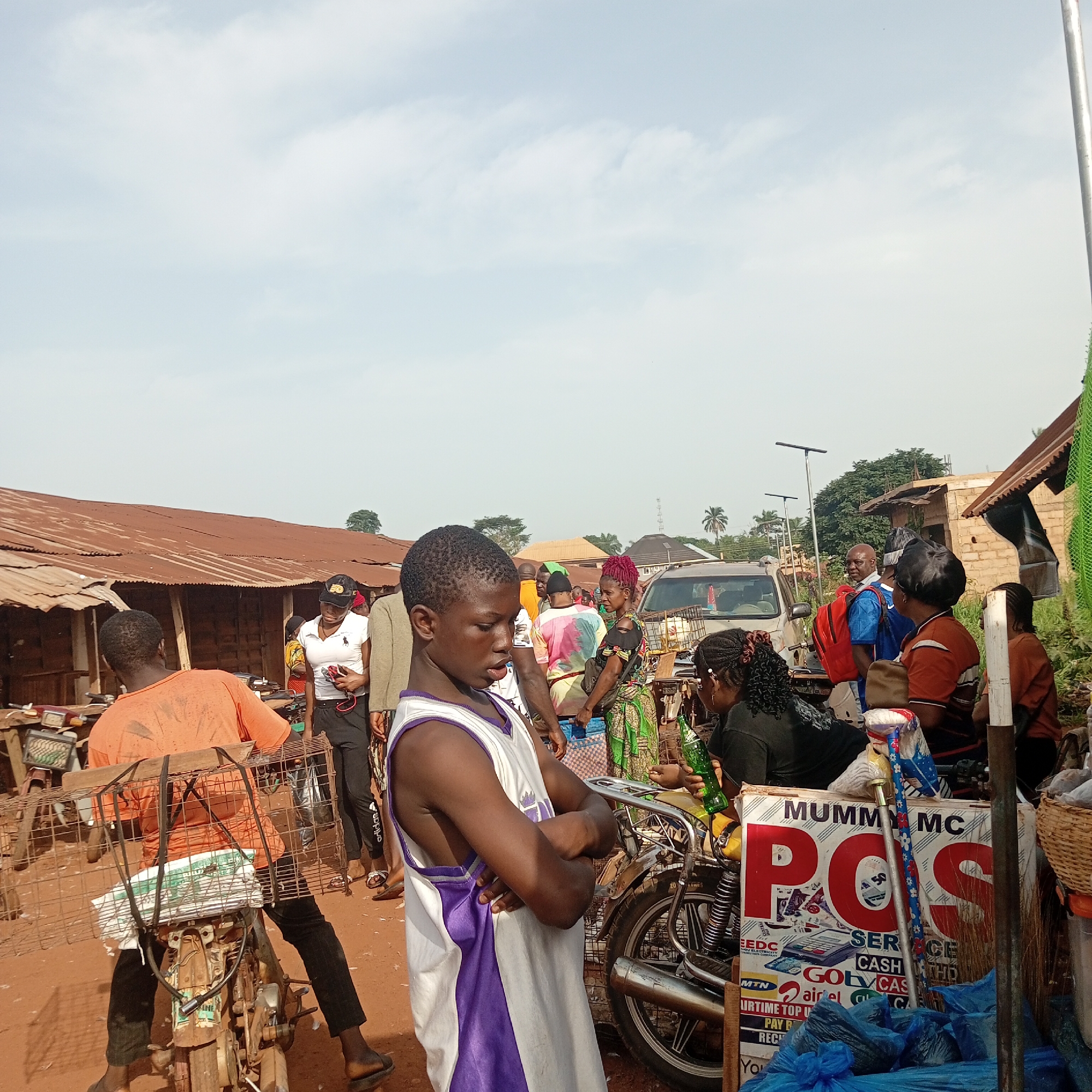 The image shows a bustling street scene in Nsukka, Nigeria. A young boy wearing a purple and white basketball jersey stands in the foreground with his arms crossed. To his right is a parked motorcycle with a sign that reads "MUMMY MC POS SERVICE CASH." A woman is seated behind the motorcycle. Another man with a cage structure is behind the boy on another motorcycle. Several people are walking around in the background, and there are buildings with corrugated metal roofs and a car in the scene. The sky is partly cloudy.