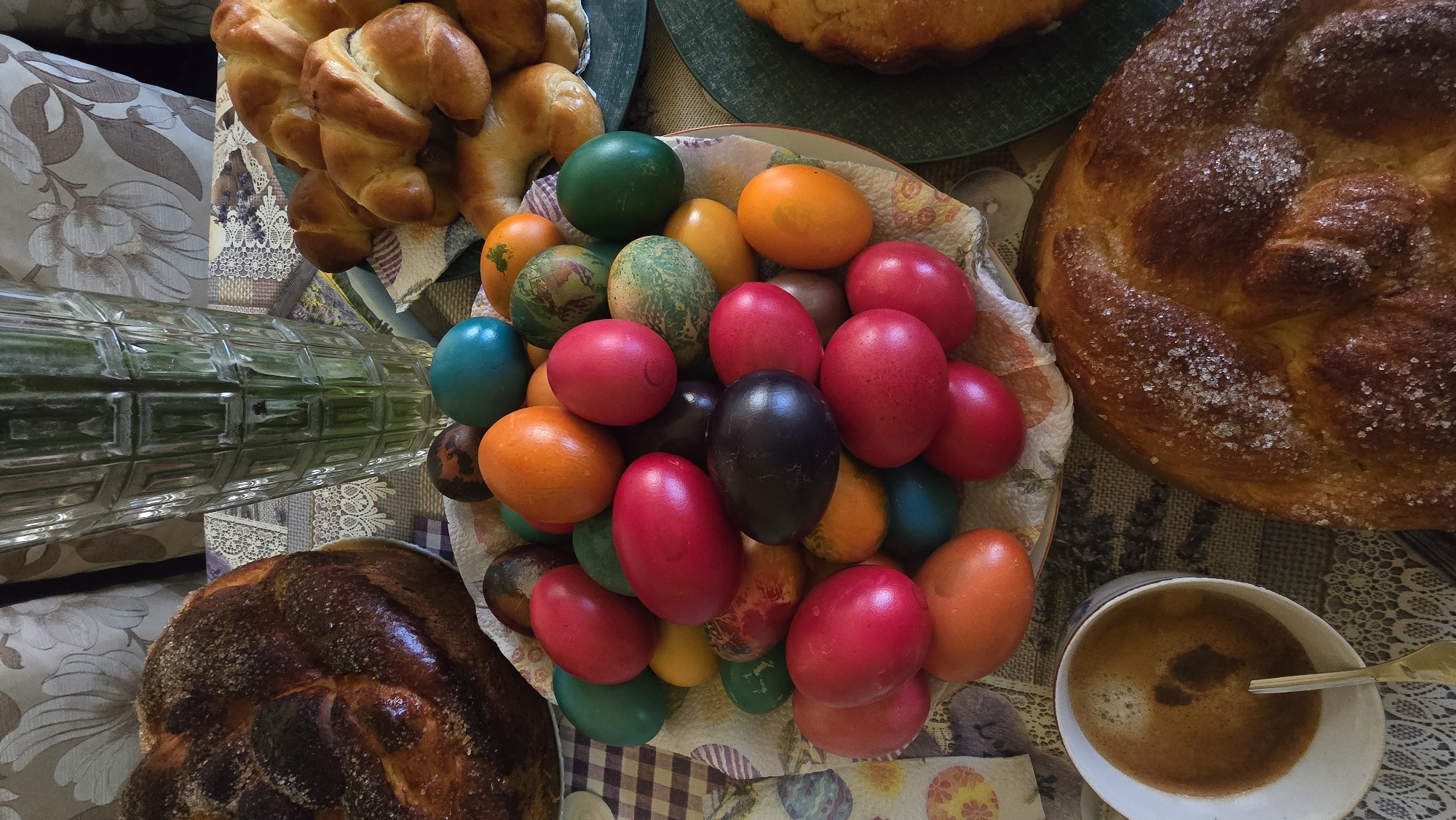 A table laden with Easter treats is the focus of this high-angle shot, possibly taken in Stara Zagora, Bulgaria. A bowl brimming with brightly colored Easter eggs, predominantly red, orange, green, and black, sits in the center, contained in a cloth-lined bowl. To the left, a glass vase is partially visible. A plate of golden-brown crescent rolls is present, and two large, glazed loaves of bread, one topped with sugar, add to the festive display. A cup of coffee with a spoon rests on the right side. The scene suggests a celebratory gathering.