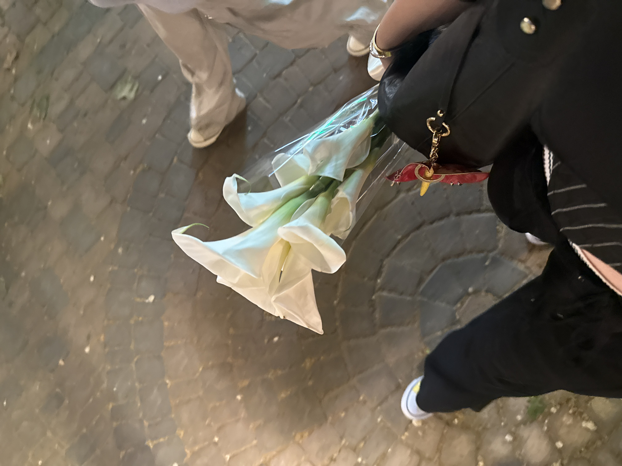 A high-angle shot shows two people on a cobblestone street. One person wears light-colored pants and white shoes. The other person wears black pants and white shoes with a yellow stripe and carries a black bag with a gold keychain. The person with the black pants is carrying a bouquet of white calla lilies in plastic wrap. The location and action cannot be determined from the image. Yerevan, Armenia is not discernible from the image.