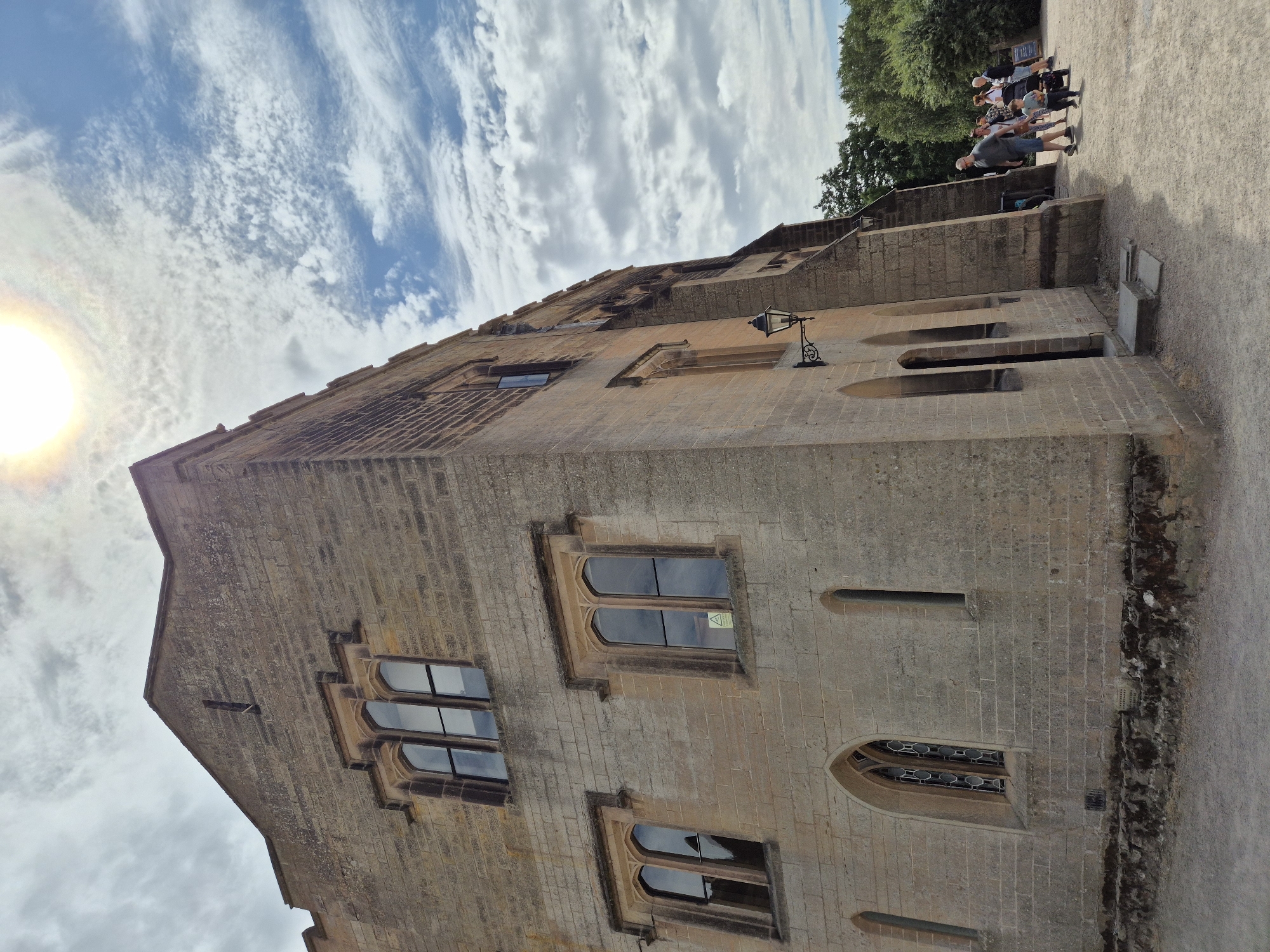 The image shows a view of the exterior of a stone building with Gothic-style windows. The building appears to be an old historical structure, possibly a castle or manor house, made of light-colored stone blocks. The sun is visible in the upper right corner of the frame, partially obscured by clouds, casting a bright light on the scene. In the lower right portion of the image, a group of people are walking on a gravel path in front of the building. A man, a woman, and a young child are prominent in the foreground, with other individuals further behind. The background consists of trees and a partly cloudy sky. The building features several arched windows, some with intricate tracery, and a single lantern mounted on the wall. The ground is covered with gravel and some patches of moss or dark vegetation at the base of the building. This scene is located at Newstead Abbey, a historic country estate located in Nottinghamshire, England, which is part of the city of Nottingham.