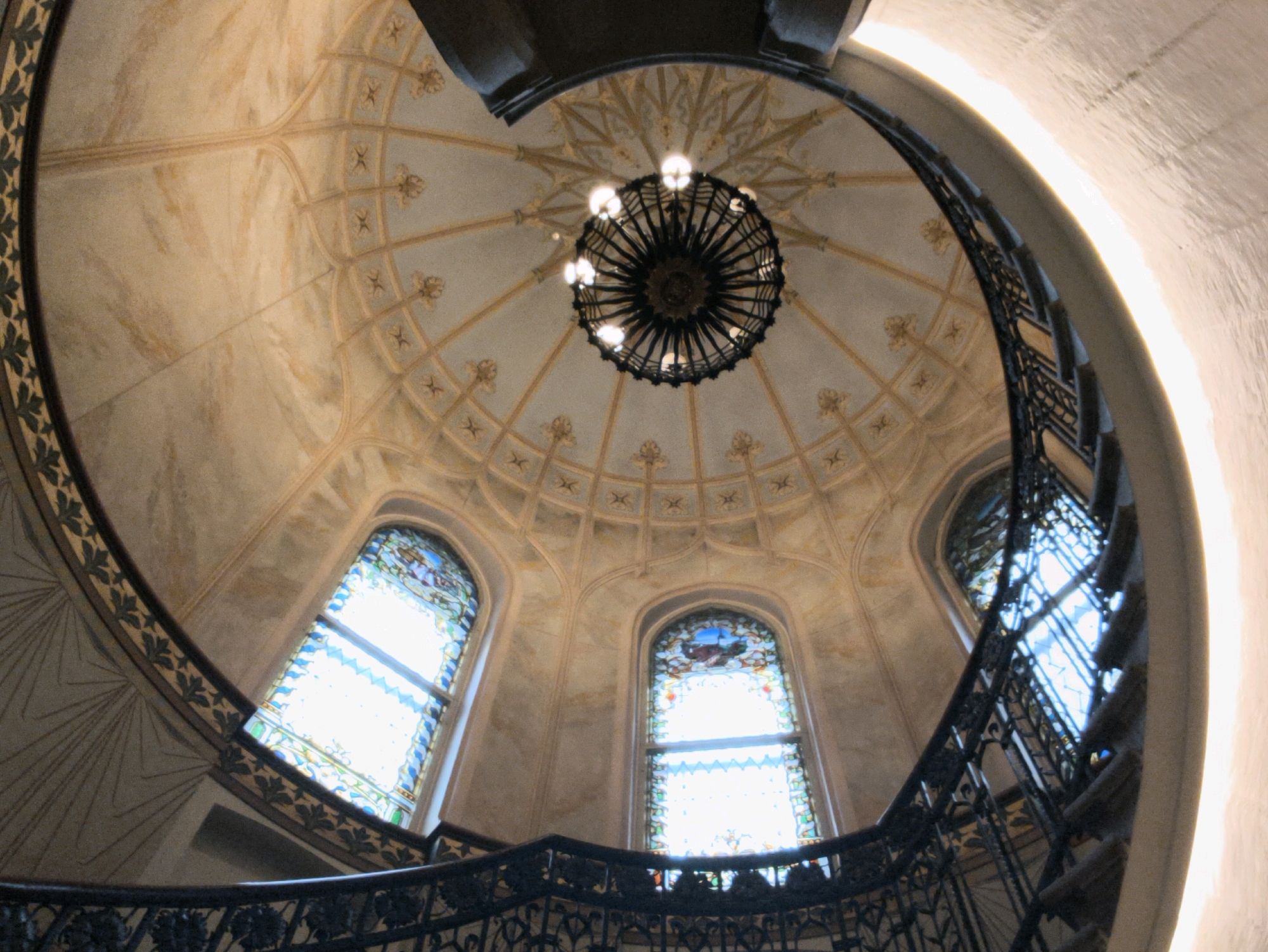 The image shows a view looking up at the rotunda of a grand staircase in Tallinn, Estonia. The rotunda features a domed ceiling with intricate golden plasterwork, radiating outwards from a large, ornate chandelier. Two arched stained-glass windows are visible on opposite sides of the rotunda, allowing natural light to filter in. A wrought-iron railing lines the curving staircase, which is visible in the foreground, spiraling upwards. The walls of the rotunda appear to be a muted, marbled tone. The overall impression is one of historical grandeur and architectural detail.