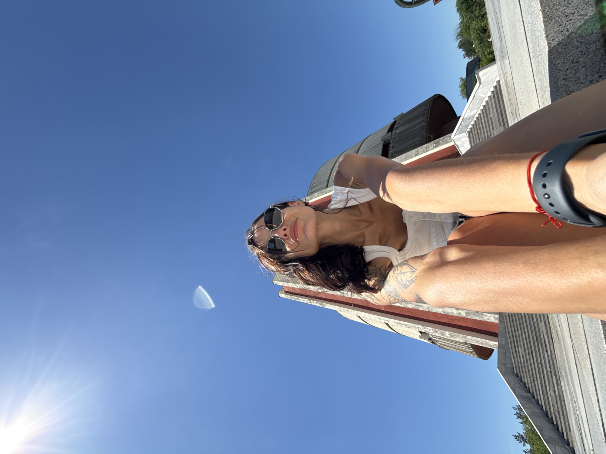 A low-angle, full-body shot captures a woman in sunglasses and a white tank top sitting on a stone structure against a bright blue sky. She is looking upwards and to her right, with her arms resting in her lap. Tattoos are visible on her arms. In the background, a large, Brutalist-style monument with red and white accents rises, featuring curved metal elements. A wide staircase with stone and concrete tiers leads up to the monument. The sun is high in the sky, casting a bright light and creating a lens flare in the upper left portion of the image. The woman is wearing a red bracelet on her left wrist. This is the Panopticon monument in Pleven, Bulgaria.