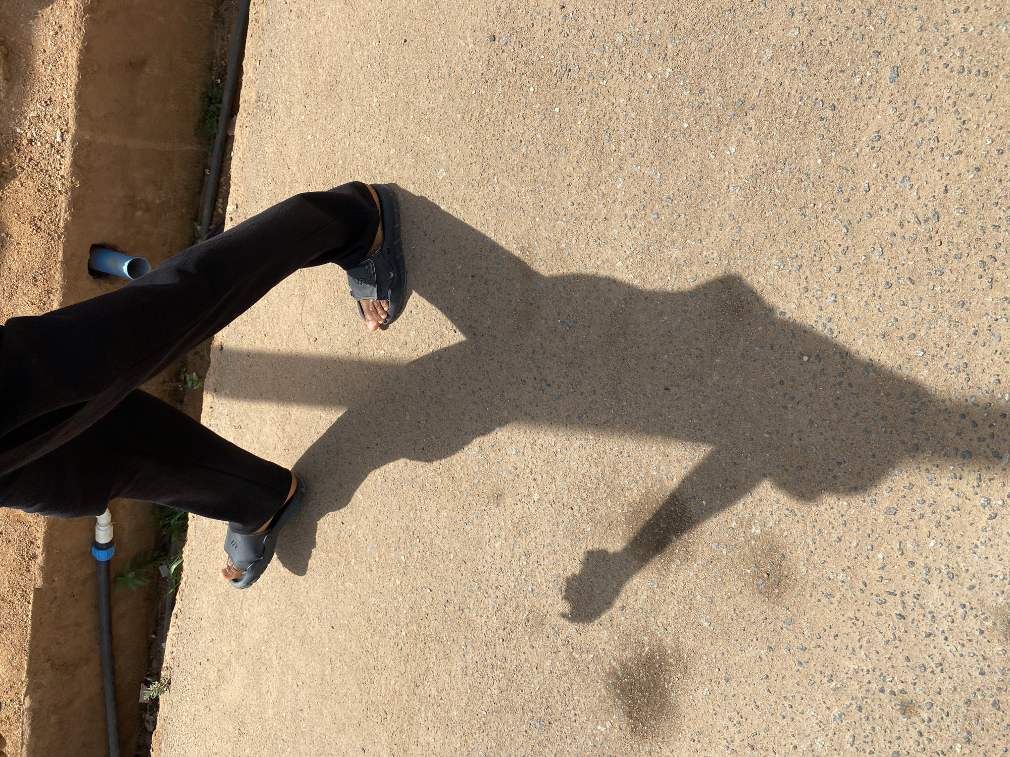 The image provides an overhead perspective of a person's lower legs and feet on a textured, light-colored concrete path. The person is wearing black pants and dark grey or black slide sandals. A prominent, elongated shadow of the person, including their upper body and arms, is cast across the concrete surface, indicating bright sunlight, likely during the mid-morning or late afternoon.
To the left of the concrete path, a shallow dirt trench is visible, containing a light blue pipe and a thinner black pipe. The black pipe features a blue and white connector near the bottom left corner. Sparse green vegetation can be seen growing within the dirt trench. The concrete surface is granular, composed of small, light-colored aggregates.
The scene depicts an outdoor setting, potentially a utility area or a pathway next to one, under clear weather conditions in Barakin Akawo, Nigeria. Faint, indistinct text or a logo is visible on the strap of the left sandal.