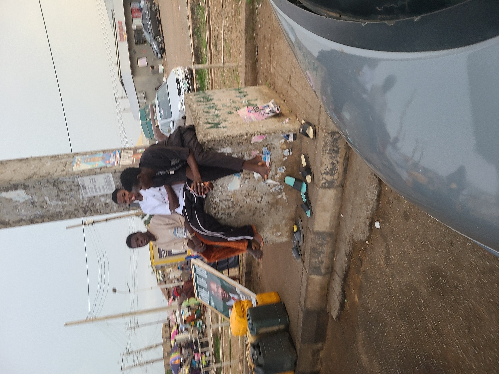 Three young men are resting on a concrete structure next to a road in Gura Top, Nigeria. One man is wearing a black shirt and pants, one is in a white shirt and black pants, and one is wearing a light brown shirt and orange pants. They appear to be sitting and lying down, relaxed. Several pairs of sandals are scattered around the concrete structure. A gray car is parked very close to the right side of the frame, partially obscuring the view. Yellow and black jerrycans are stacked near a sign propped up on the ground. Power lines are visible in the background and there are cars and buildings further in the distance. The ground appears to be asphalt and has some visible debris.
