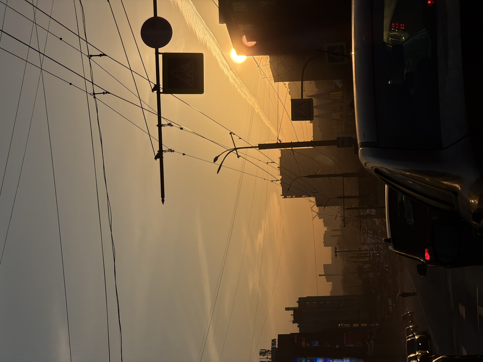 The image depicts an urban scene in Izhevsk, Russia, during what appears to be a sunset or sunrise. The sky is a warm, golden hue, with a contrail visible behind the sun. Overhead wires crisscross the sky. Street signs are attached to poles, with pedestrian crossing signs visible. Silhouetted buildings line the street. Cars are parked on the street, with a black car taking up a significant portion of the right side of the image. The photo is rotated counterclockwise.