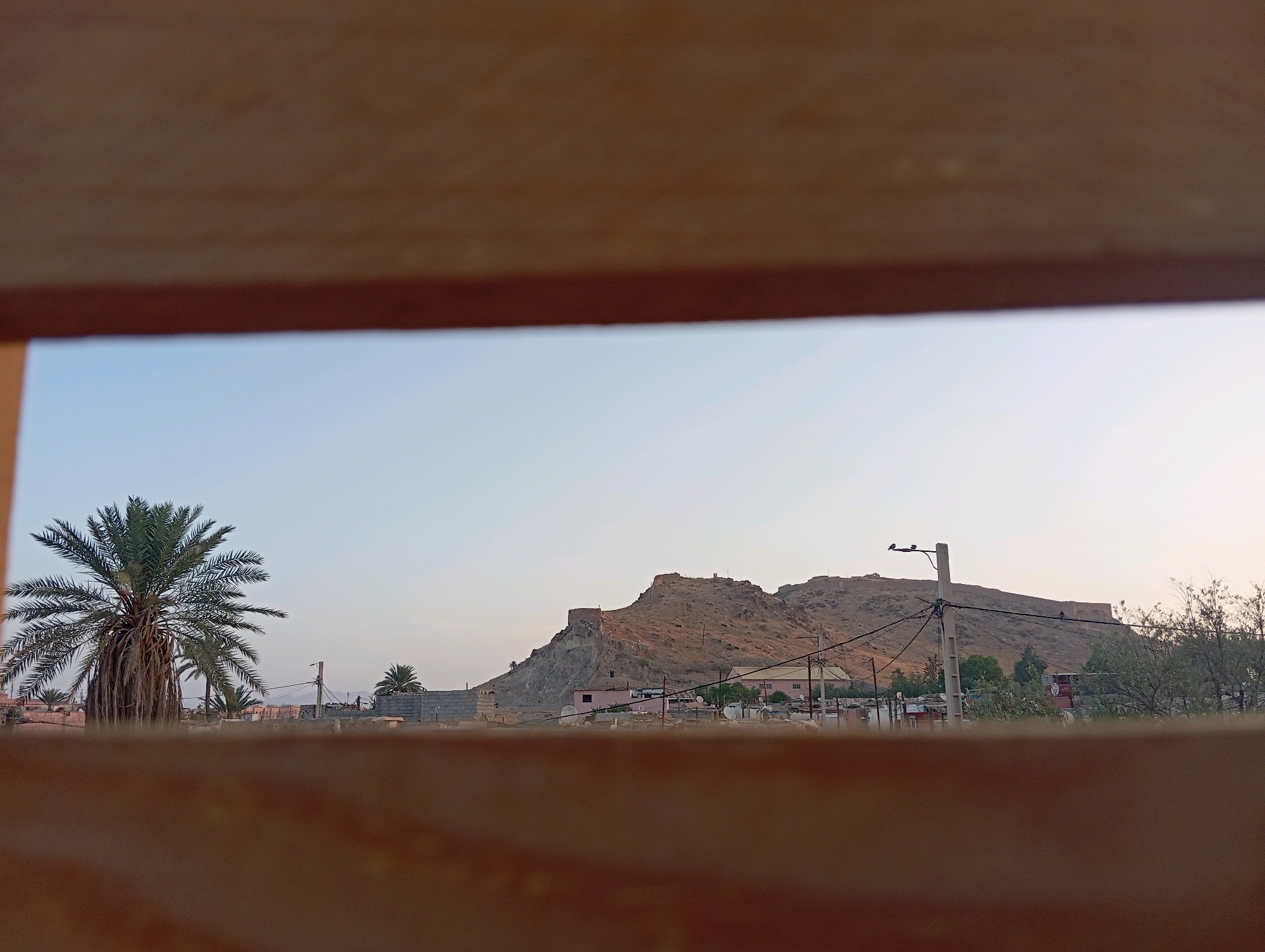The image depicts a view through a wooden frame, revealing a landscape with a fortified hilltop in the distance. A prominent date palm tree stands on the left side of the frame, with its fronds extending upwards against a pale blue sky. In the middle ground, several buildings, some appearing to be made of concrete blocks and others with pink facades, are visible. Electrical poles and wires crisscross the scene, and trees and other vegetation are scattered throughout. The fortified hilltop, likely an ancient kasbah or ruin, commands the horizon. While the image is said to be located in Marrakesh, Morocco, no specific landmarks within the city are immediately identifiable. The scene appears to be a rural or semi-urban setting, possibly on the outskirts of the city, characterized by its arid environment and traditional architecture. There are no people or animals clearly visible in the image. The overall impression is one of a quiet, historic landscape bathed in the soft light of either early morning or late afternoon.