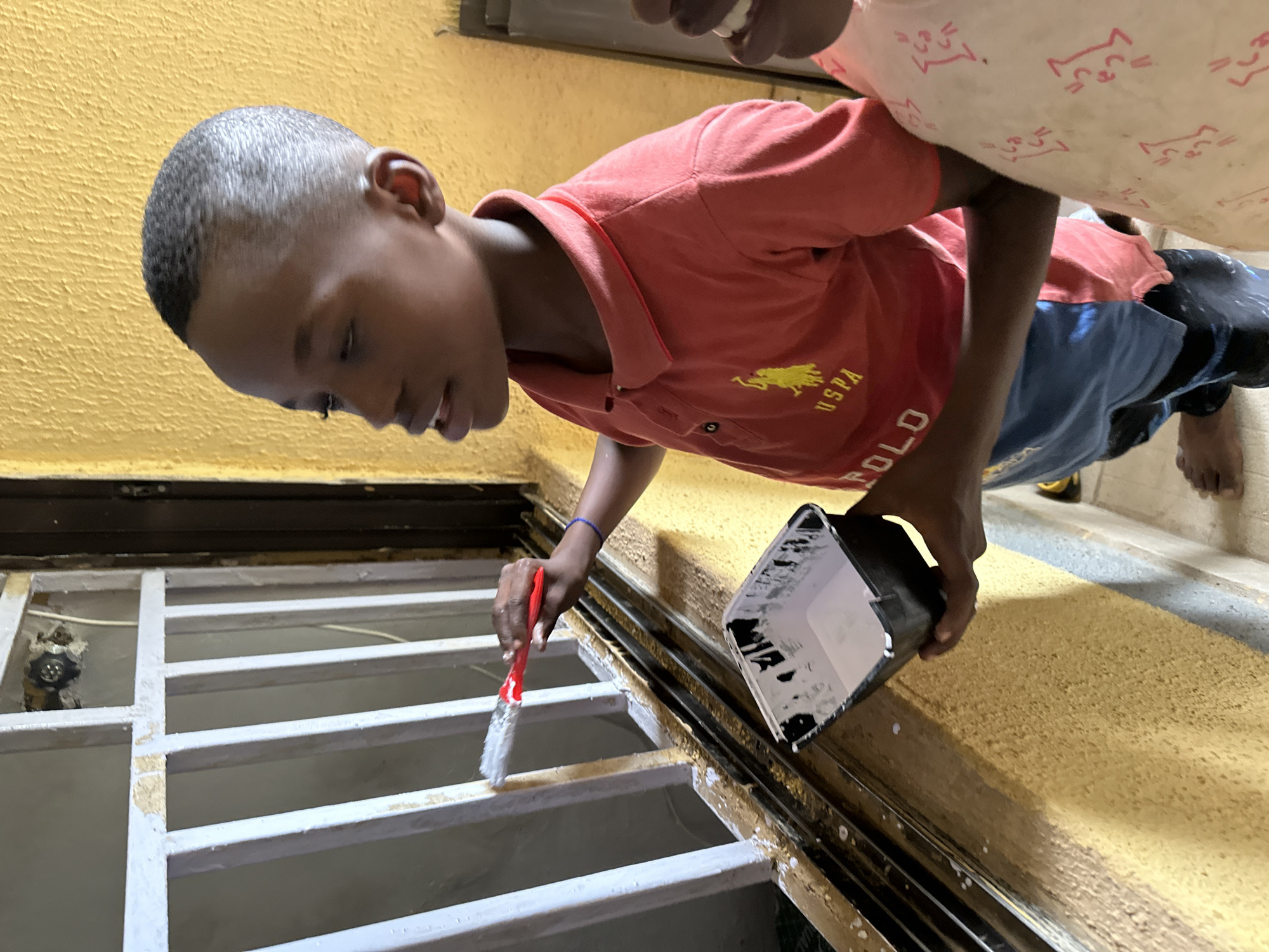 In Narayi, Nigeria, a young boy with a close-cropped haircut is painting a white metal grill with a red-handled brush. He is wearing a red polo shirt with a yellow USPA logo and blue shorts. He holds a black container of white paint in his left hand. The wall behind him is yellow and textured. The scene appears to be outdoors, possibly on a balcony or porch. Another person's arm and shoulder, wearing a light-colored garment with a pink pattern, are visible on the right side of the frame.