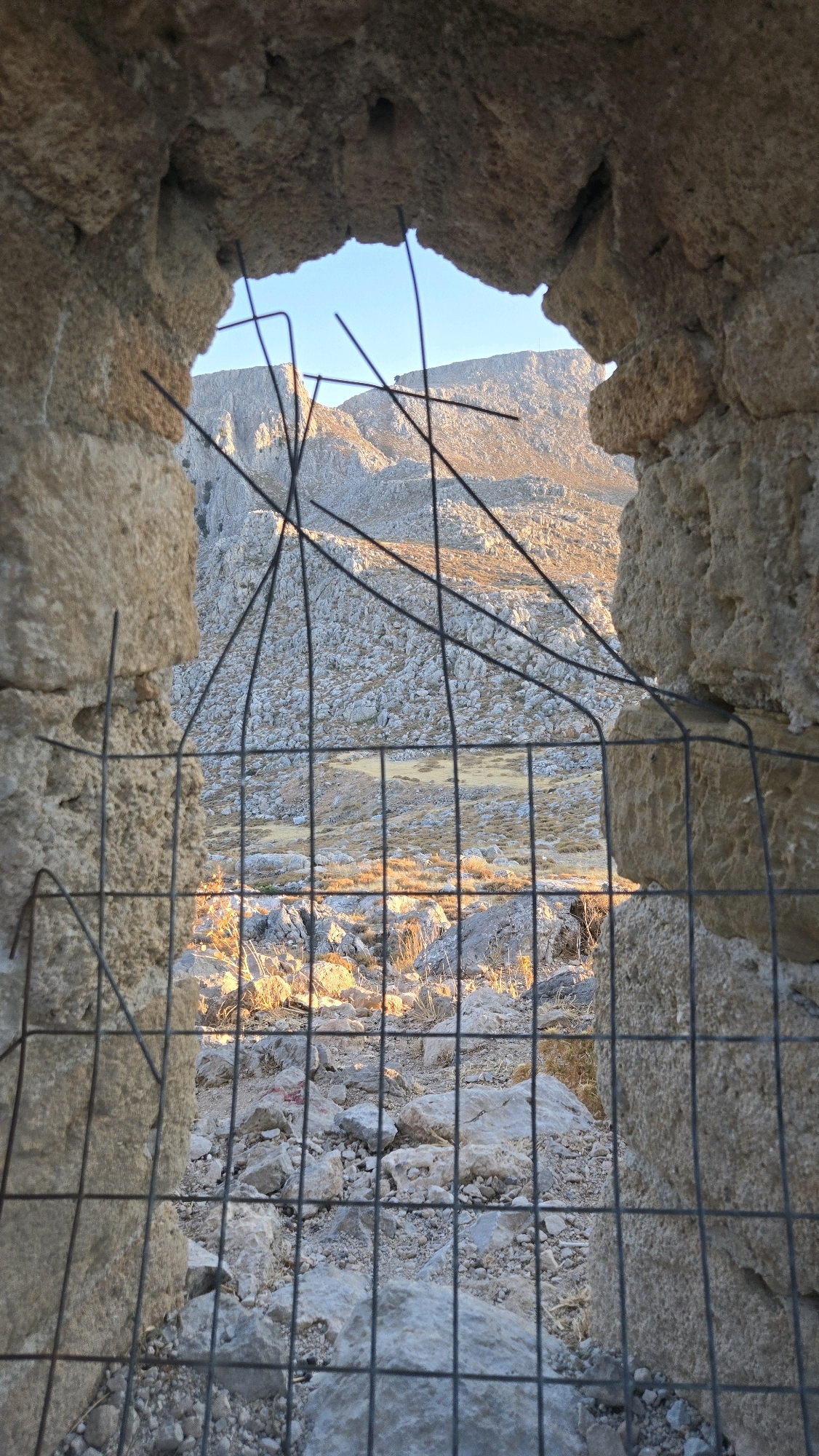 This image captures a view through a rough opening in a stone structure, possibly a ruin or a historical building, in Archangelos, Greece. The opening is partially obscured by a wire mesh fence. Through the aperture, a rugged, rocky landscape with sparse vegetation is visible under a clear blue sky. The sunlight, likely from a low angle, casts warm, golden hues on the distant mountains and terrain, creating strong shadows and highlighting the textures of the rocks. The foreground is dominated by the rough-hewn stone of the opening and the metallic grid of the fence, which is made of dark, thin wire forming a crisscross pattern. The rocks in the immediate foreground are also rugged and appear to be part of the same natural or constructed environment. The scene conveys a sense of desolation and natural beauty, with the man-made barrier of the fence adding a layer of human presence or intervention.