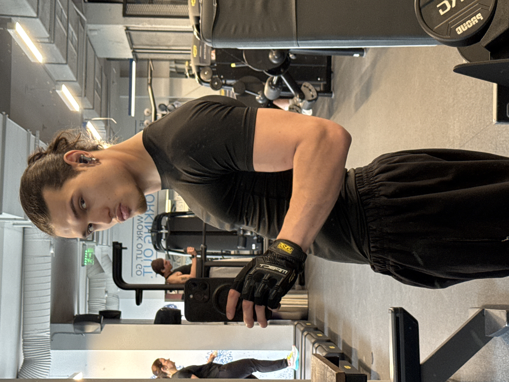 This image captures a young man taking a selfie in a gym, likely located in Sofia, Bulgaria. The man, with dark hair tied back, is wearing a black short-sleeved t-shirt and black workout pants. He has black earbuds in his left ear and is holding a black smartphone with his right hand to take the picture. His right hand is covered by a black, fingerless workout glove featuring yellow branding that includes the word "IMPACT" and a distinctive "M" shaped logo, possibly "MECHANIXX".
The background reveals a typical gym setting with various exercise equipment, including weight machines and racks. The space is illuminated by fluorescent strip lights on the ceiling. A large mirror on the wall reflects the gym environment, other gym-goers, and reversed text. Reflected on a sign in the mirror is the text "ORKING OUT" and "eo", which is likely a reversed "WORKING OUT.co". A weight plate visible in the upper background also shows reversed text, appearing as "DUOBD".
In the mirror's reflection, another individual, possibly the same man from a different angle, is seen working out with weights on a bench. Further down, another person is partially visible lying on the floor, possibly stretching. The gym floor is a light grey, and white corrugated air ducts are noticeable along the walls and ceiling. The image depicts an active gym session, with the man posing for a personal photo.