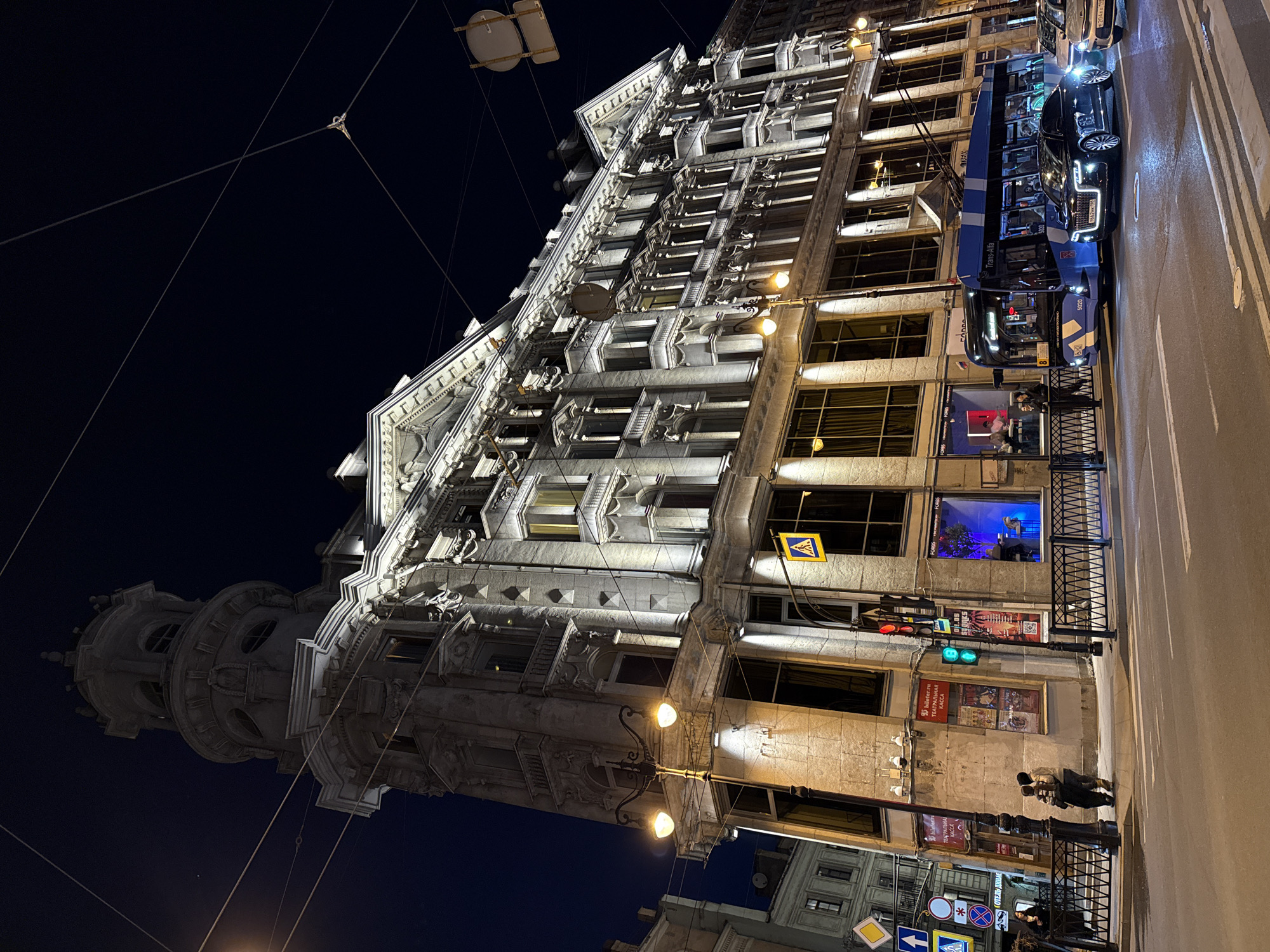 The image is a low-angle shot of a building, probably in Saint Petersburg, Russia, taken at night. The building is brightly lit, with detailed architectural features, including a round tower on the left and numerous windows, many of which are dark, along its length. There is a trolley bus with the words "Trans-Alfa" written on the side and a number "5020" on the right of the building, parked on the street. A black car is next to the bus. The street is paved, with traffic signs and signal lights. There are two people walking on the sidewalk. There are street lights and overhead wires visible against the dark night sky. Banners and posters are visible on the side of the building, as well as a sign with theatre advertising (theatre ticket box office).