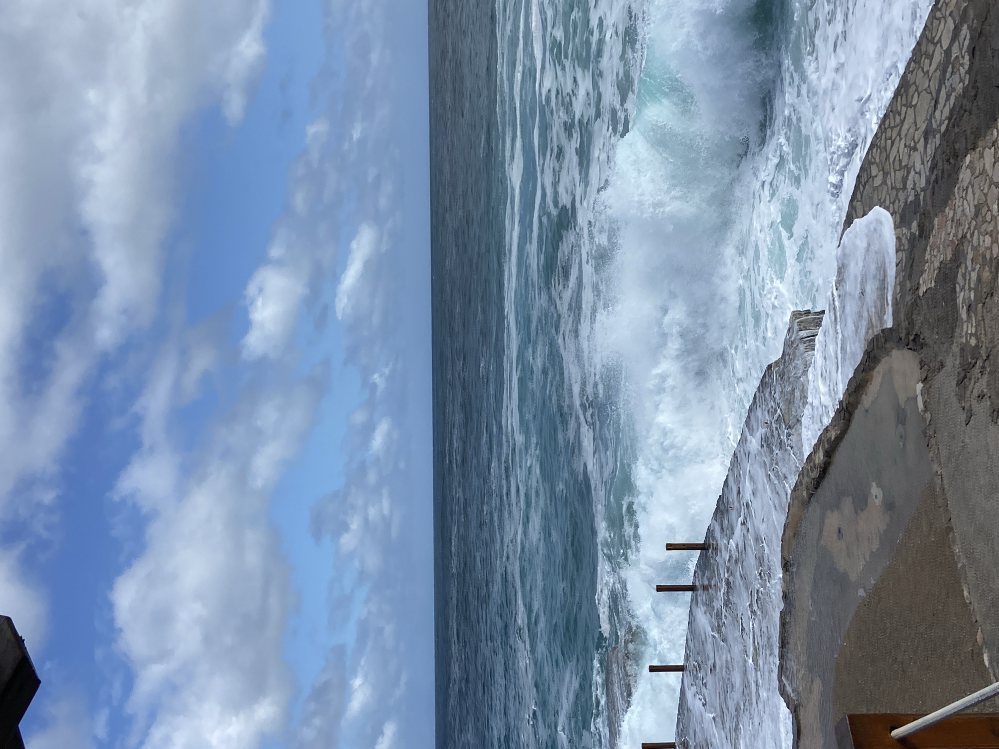 The image shows a view of the ocean crashing against a rocky structure. The sky, with blue and white clouds, is visible on the left side. The ocean occupies the central and right portions of the image, and the foamy surf of a breaking wave is prominent. On the far right, a portion of what appears to be a man-made structure, possibly a concrete or stone wall, is visible, indicating a constructed element near the shoreline. There are wooden planks embedded on the wall. The image appears to be taken at the city of Prijevor, Montenegro.