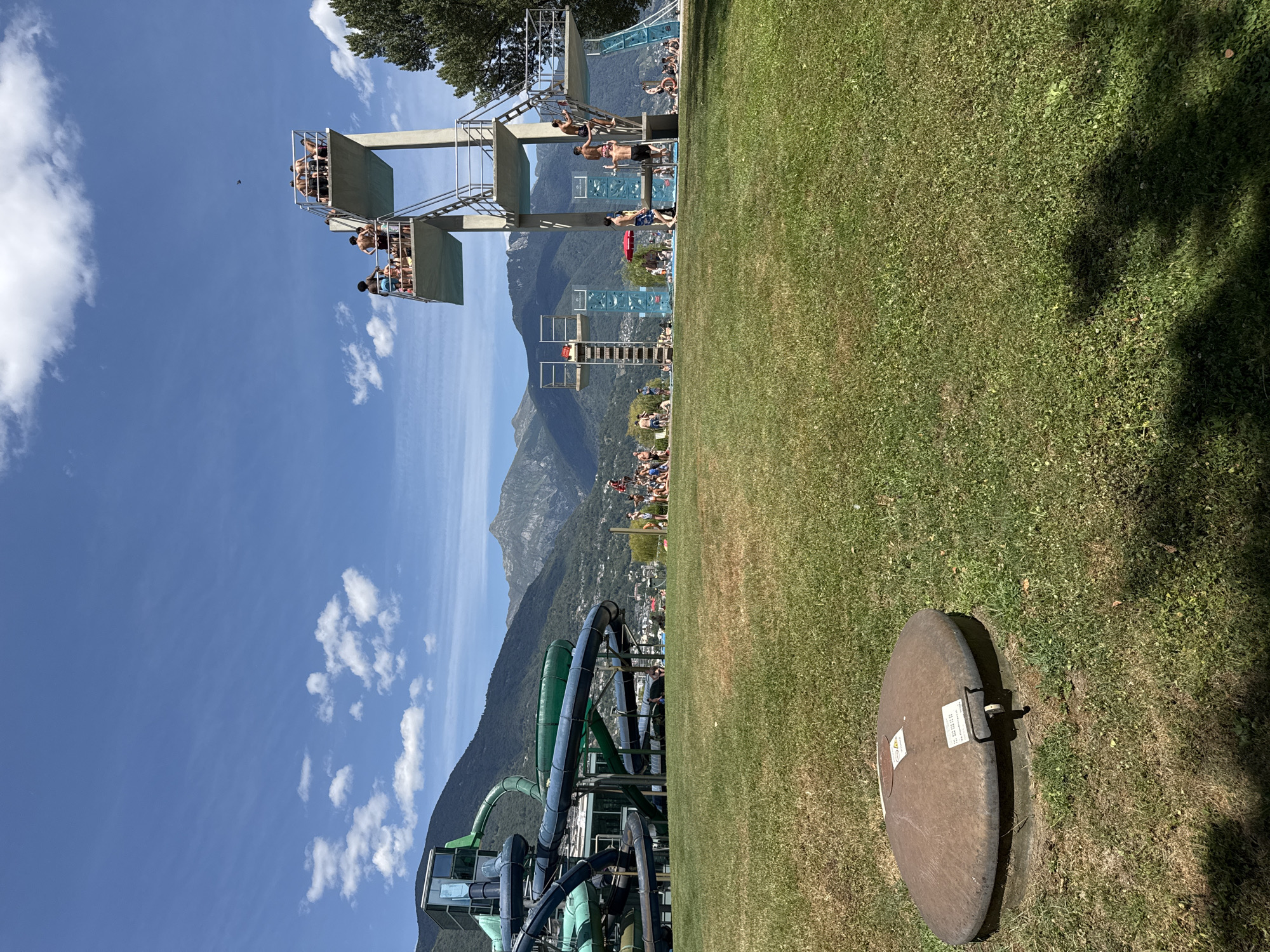 In Locarno, Switzerland, this image captures a vibrant summer day at an outdoor water park, likely Lido Locarno, with the majestic Alps forming a scenic backdrop. The foreground is dominated by a lush green lawn, with a large, circular metal manhole cover with two white labels near the bottom left. Beyond the lawn, a complex of blue and green water slides snakes across the frame, their colorful tubes contrasting with the natural landscape. To the right, a multi-level diving platform stands tall, with several individuals in swimwear on its platforms and stairs, preparing to jump. Other park-goers are visible throughout the scene, some relaxing on the grass, others near the water slides or diving boards, enjoying the sunny weather. The sky above is a brilliant blue, dotted with scattered white clouds, and the mountains in the distance are covered in greenery, with some rocky peaks visible. The overall atmosphere is one of leisure and recreation under a clear, sunny sky.