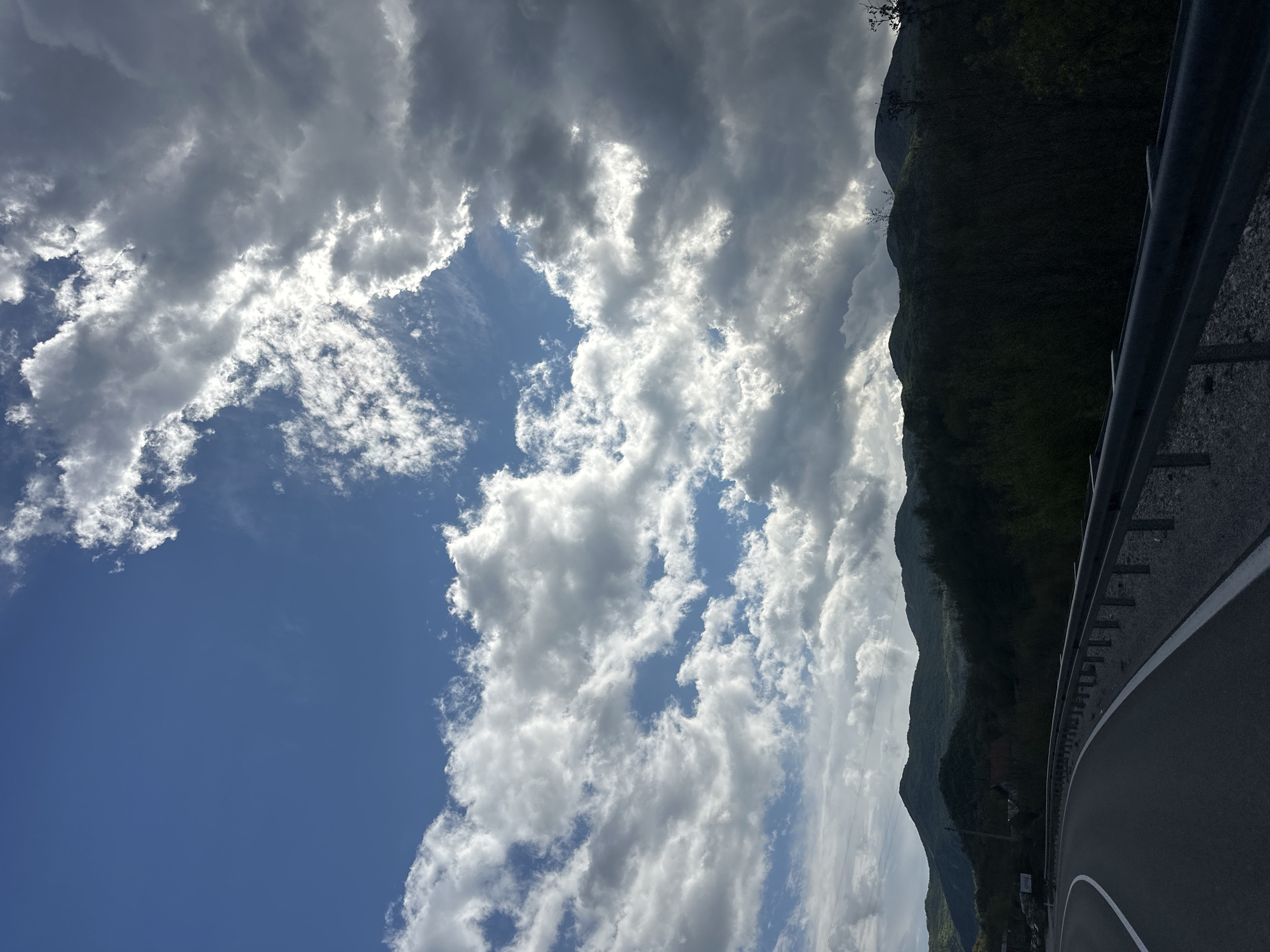 The image captures a majestic mountainous landscape, likely in Očinići, Montenegro, under a dramatic sky. The scene is dominated by a vast expanse of dynamic clouds and a towering, forested mountain range.
The sky, occupying the left and upper portions of the frame, is filled with a mix of bright white cumulus and darker stratocumulus clouds. Patches of clear, deep blue sky are visible between the cloud formations, particularly in the lower left. The sun, though not directly visible, brightly illuminates the edges and undersides of many clouds, creating a luminous effect and suggesting a bright, partially cloudy day.
On the right side, a steep, densely vegetated mountain rises sharply, covered in dark green foliage, indicative of a rugged, natural environment. Its peak extends beyond the top of the frame. In the foreground, a paved road curves from the bottom right towards the middle right, alongside the mountain. A robust metal guardrail runs parallel to the road, providing a sense of scale and safety for the winding path. White road markings are discernible on the dark asphalt surface.
The time of day appears to be daylight, with strong natural illumination. No people, vehicles, or animals are visible, emphasizing the grandeur and solitude of the natural environment. A small, white rectangular sign is present near the roadside at the base of the mountain, but its text is indecipherable. The overall impression is one of serene yet imposing natural beauty.