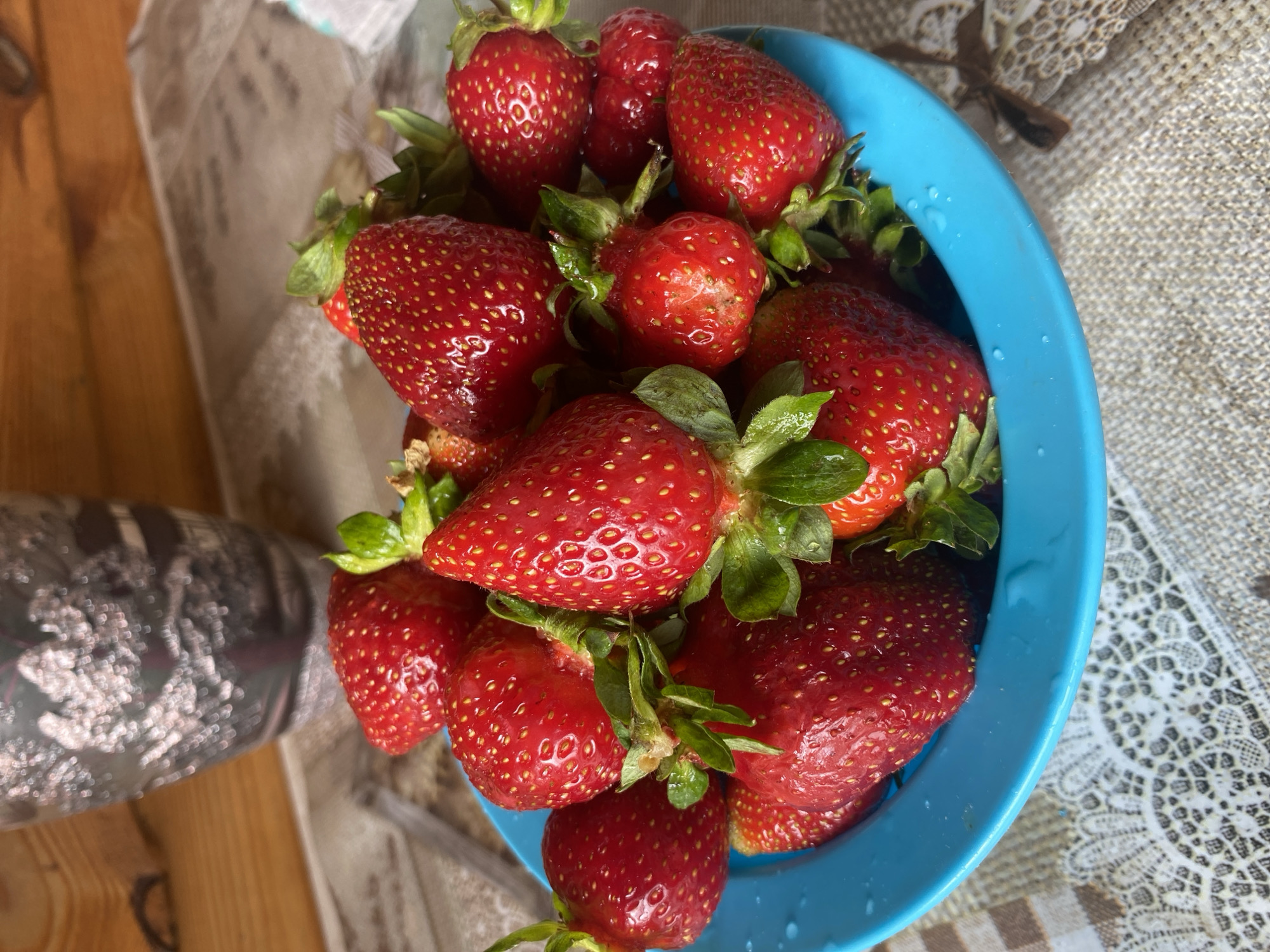 A close-up, overhead shot shows a blue bowl overflowing with ripe, red strawberries. The strawberries are plump and have visible seeds. Some have green stems and leaves attached. The bowl is placed on a patterned tablecloth. In the background, a decorative silver vase is visible against a wooden surface. The lighting suggests an indoor setting, possibly a kitchen or dining area. The image does not provide any information about the city of Chaikovsky, Russia.