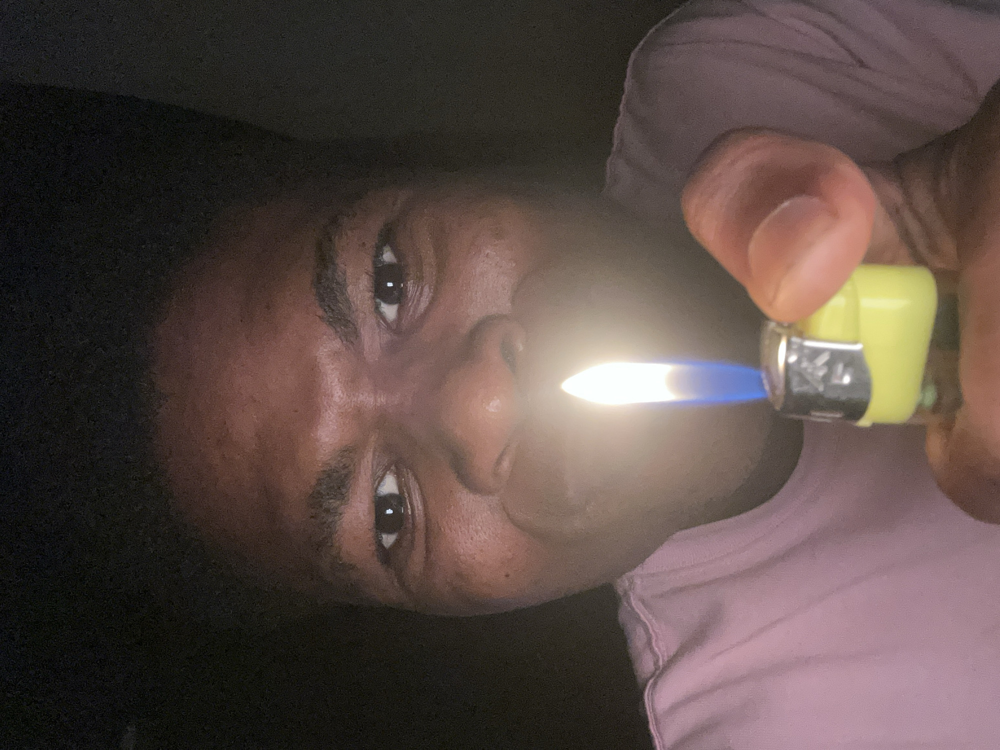 The image shows a close-up of a person holding a lit lighter, with the flame illuminating their face. The person has dark skin and is wearing a pink t-shirt. The background is dark, suggesting the photo was taken at night or in a dimly lit environment. The location of Ihiagwa, Nigeria is not discernible from the visual information provided in the image.