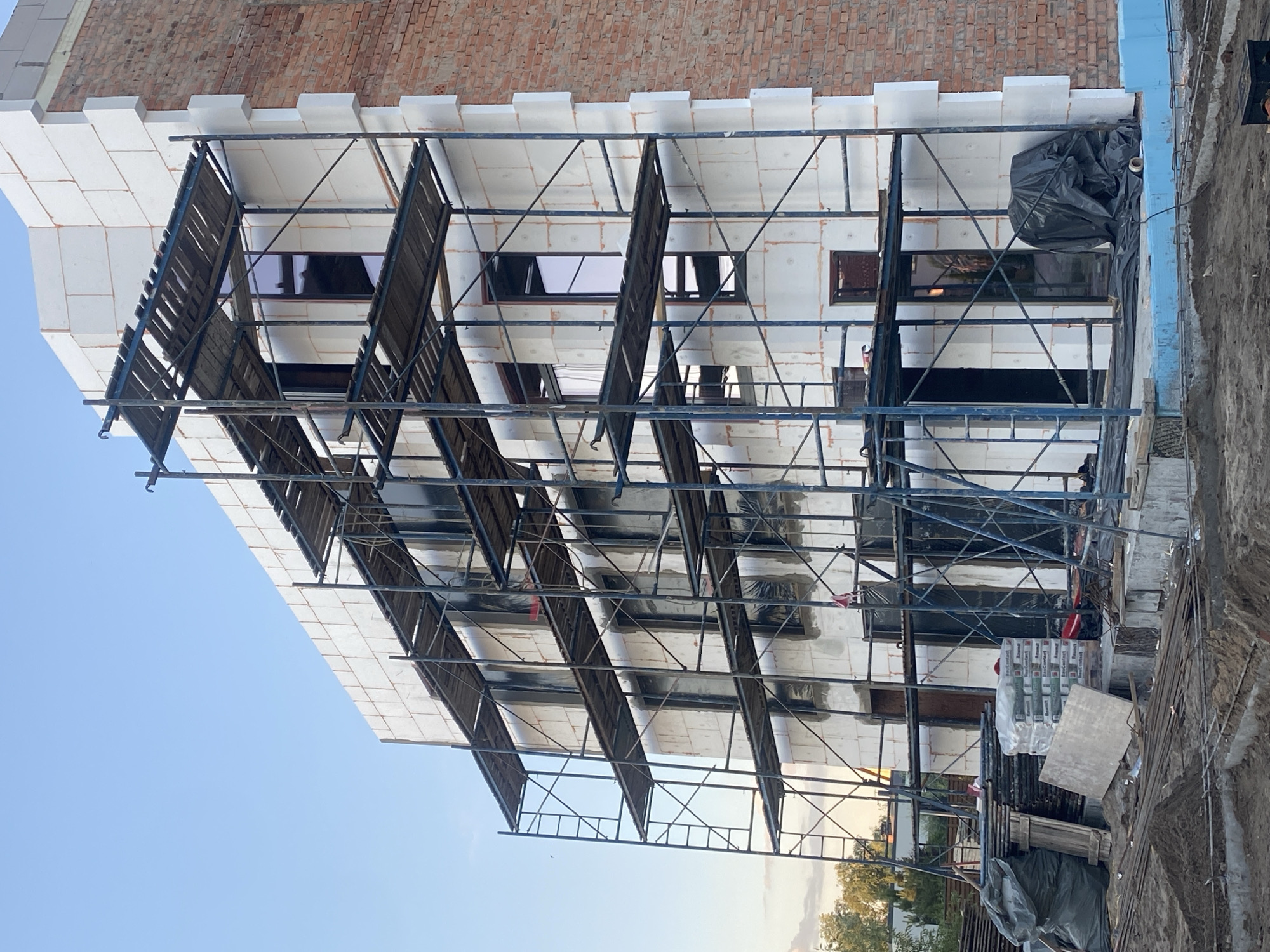 The image shows the exterior of a multi-story building under construction in Novi Petrivtsi, Ukraine. The building features scaffolding on its facade, which is covered in white insulation panels. A section of the building's exterior is also visible with exposed red brick. Several windows are present on the building, some covered with dark material. Construction materials, including bags of what appear to be cement or insulation, lumber, and metal rebar, are scattered around the base of the building. The ground is a mix of dirt and exposed concrete foundations. In the background, trees and a clear blue sky are visible. There are no people visible in the image.
