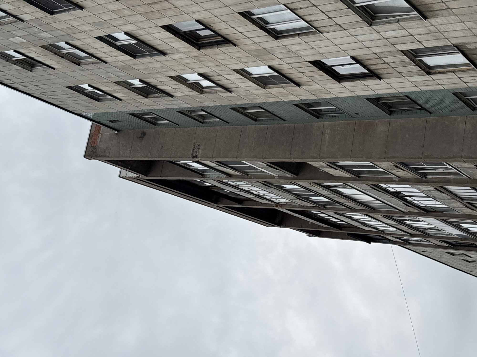 The image depicts a close-up, low-angle view of the exterior of a Brutalist-style apartment building in Saint Petersburg, Russia. The building's facade is predominantly composed of concrete tiles and rectangular windows, some of which appear to be open or have visible interior structures. There is a protruding concrete balcony or structural element that casts a shadow. The overall aesthetic is stark and utilitarian, characteristic of Soviet-era architecture. The sky is overcast and grey, suggesting a cloudy day. No people are visible in the frame, and the scene appears quiet and possibly uninhabited, emphasizing the architectural subject. The time of day is indeterminate due to the uniform lighting conditions. The image is rotated 90 degrees counterclockwise, so the building appears to be upside down from a typical perspective.
