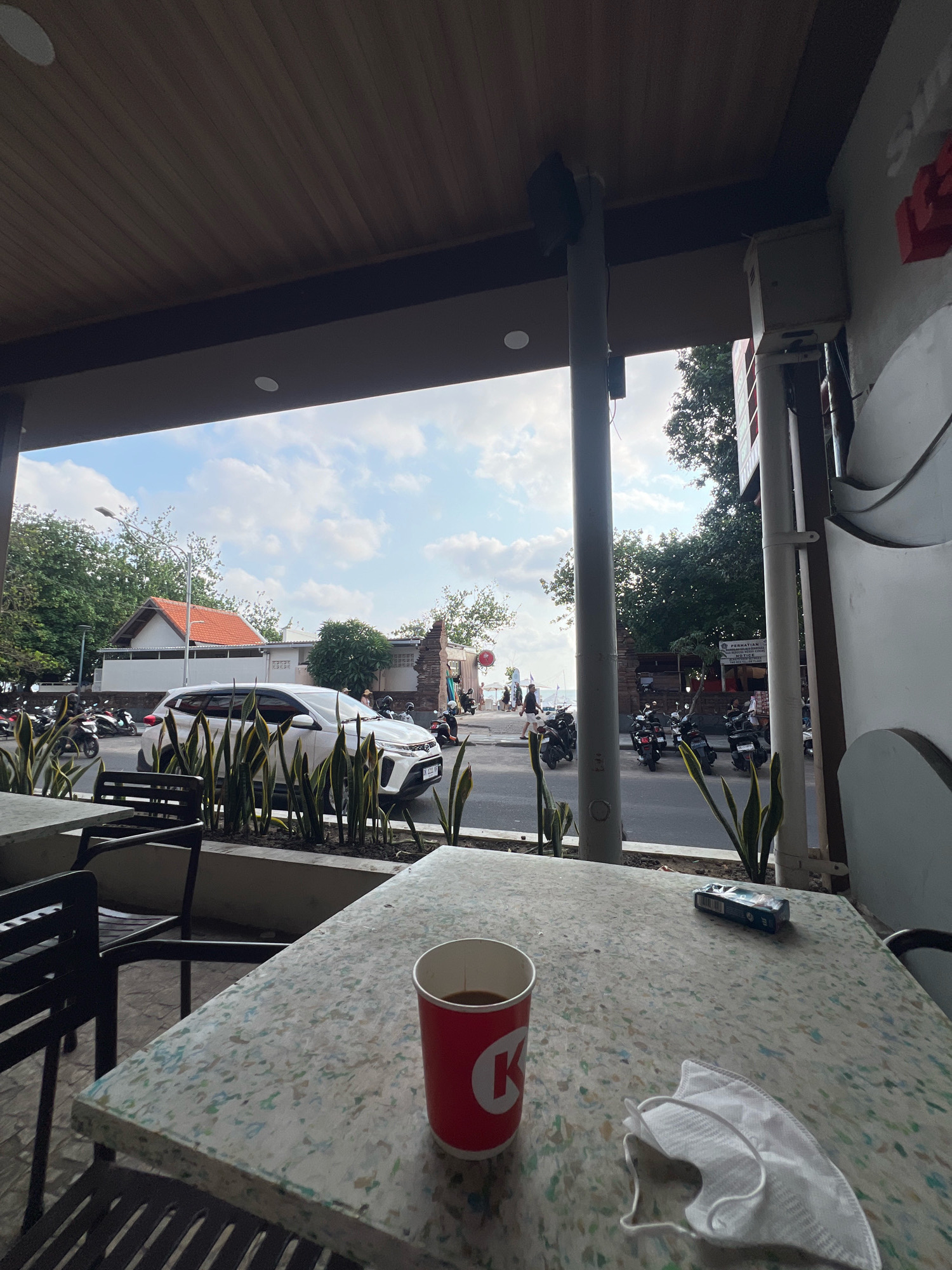 The image depicts a street-side view in Kuta, Indonesia, looking out from what appears to be a covered seating area, likely a cafe or restaurant. In the foreground, a speckled, light-colored table holds a red disposable cup with a white logo (partially visible as a 'K'), a packet of cigarettes, and a white disposable face mask. Two black chairs with slatted seats and backs are positioned around the table.
Beyond the seating area, a busy street is visible. Several motorcycles are parked along the roadside, and a white SUV is in motion, driving away from the viewer. A sidewalk runs alongside the street, with lush green plants in planters separating the seating area from the road. Trees line the street, and a white building with a terracotta-tiled roof is prominent. In the background, a glimpse of the ocean can be seen under a cloudy sky. The architecture suggests a tropical setting, consistent with Kuta. The overall scene captures a moment of daily life in a coastal town, with elements of both leisure and transit.