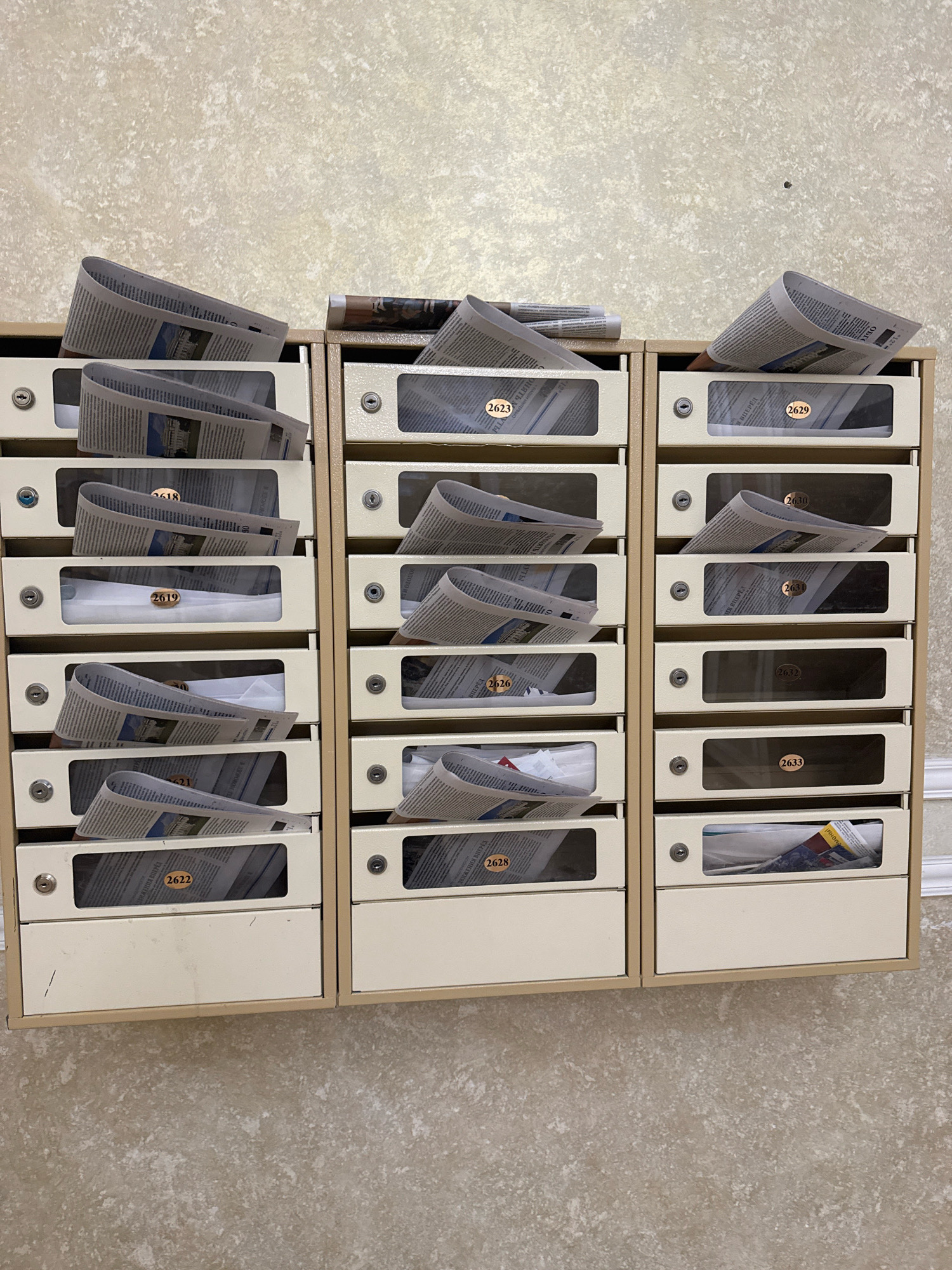 The image depicts a bank of mailboxes, likely in a residential building in Saint Petersburg, Russia. There are three columns of mailboxes, each with multiple compartments. Many of the compartments are filled with newspapers, some of which are folded and sticking out. Some compartments have numbers on them, such as 2623, 2629, 2618, 2619, 2620, 2631, 2632, 2633, 2626, 2628, and 2622. The mailboxes are a neutral beige color and are mounted on a wall with a textured, light-colored finish. The scene is well-lit, suggesting it was taken indoors. There are no people visible in the image.