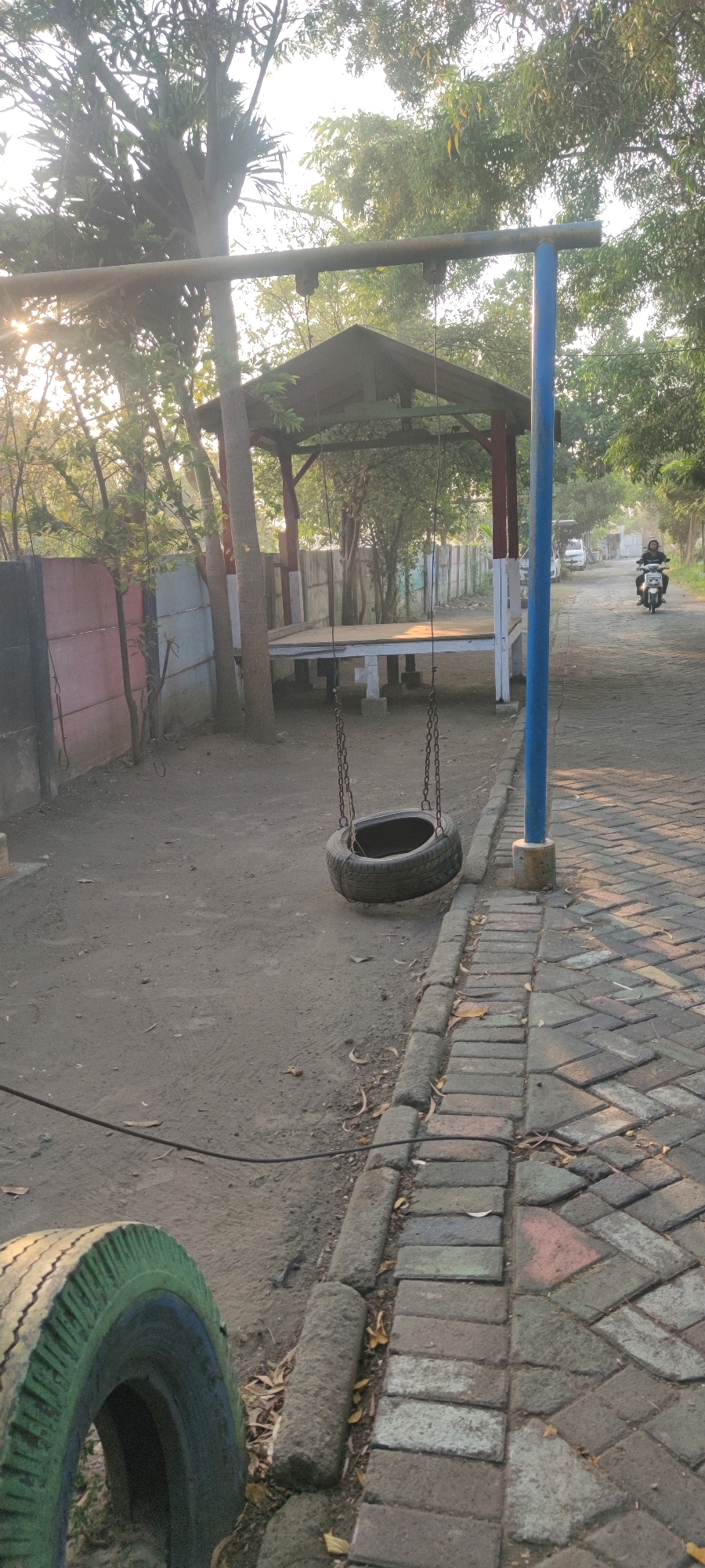 In Tebel, Indonesia, a tire swing hangs from a metal frame in a park setting. The swing, made from a black tire, is suspended by two metal chains. To the left of the swing, a wall painted in sections of pink and white borders a dirt ground. Behind the wall are trees. In the background, a covered pavilion with a thatched roof stands on a raised platform. A paved walkway with a brick pattern runs alongside the dirt area, and a blue pole is planted near the walkway. Further down the walkway, a person rides a motorcycle. The environment appears to be outdoors, with sunlight filtering through trees, casting shadows on the ground.