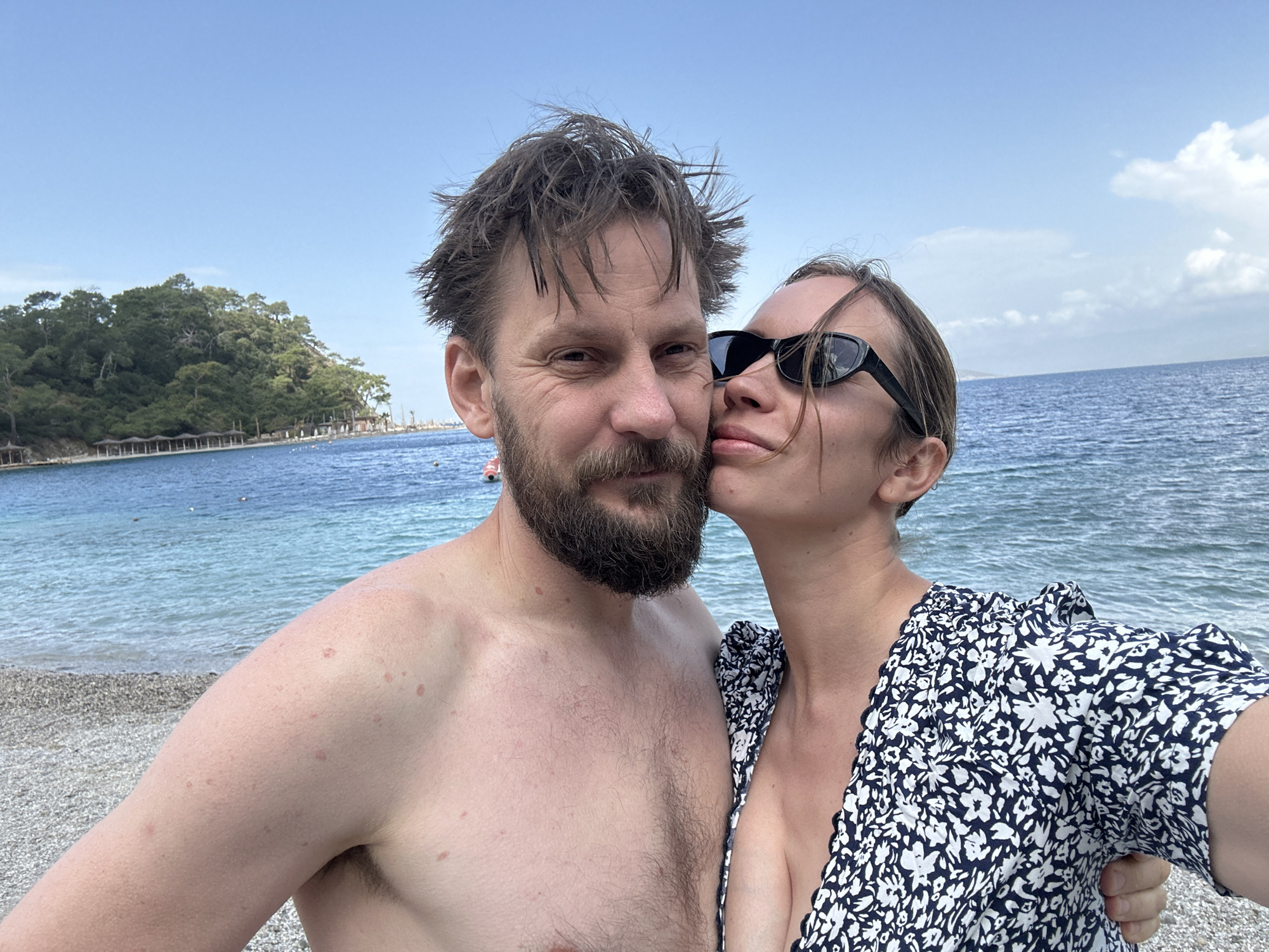 A photo depicts a couple on a beach near Fethiye, Turkey. The man is bare-chested with dark hair and a beard. The woman is wearing a black and white floral dress and black sunglasses. She is kissing the man's cheek. The backdrop features a pebbly beach and a sea with tree-covered islands or peninsulas in the distance. The sky is partly cloudy.
