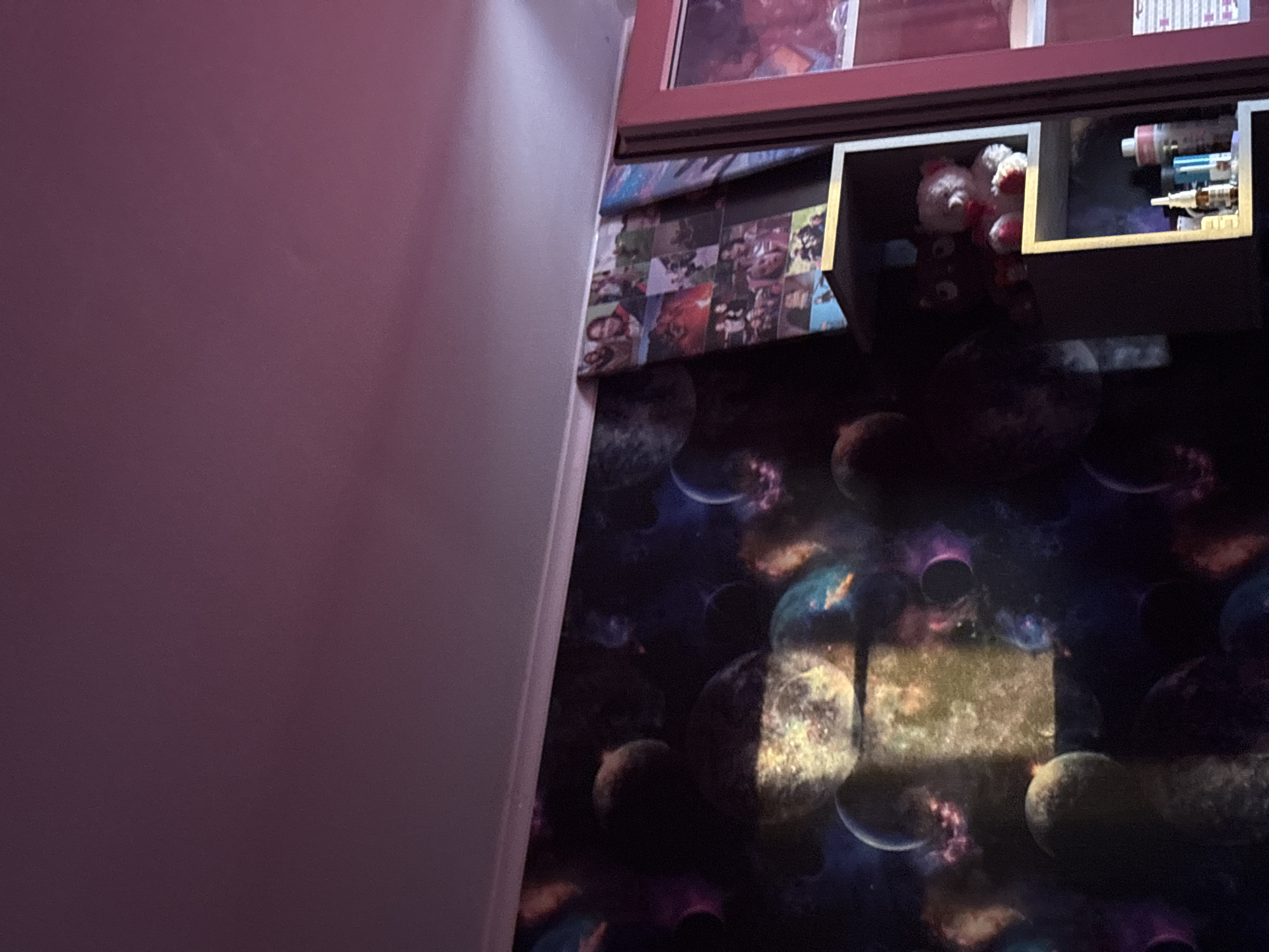 The image depicts a section of a room, focusing on a corner with a shelf unit and adjacent wall. The wall is painted in a solid, light shade of purple. The shelf unit is composed of multiple compartments and appears to be made of dark wood or a similar material. The shelf unit has multiple levels with boxes holding various items, including what appears to be plush toys and medication. The wall behind the shelf is decorated with a space-themed wallpaper featuring celestial bodies and nebulae. Additional printed materials are affixed to the wall between the shelf unit and the wallpaper.
