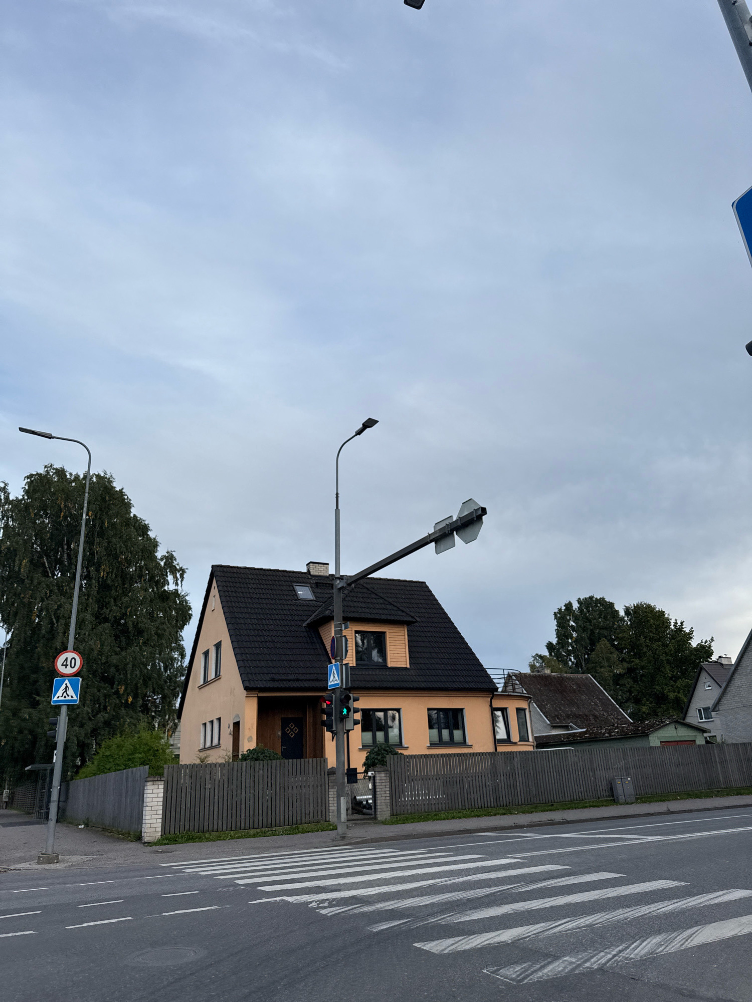 The image shows a street scene in Tallinn, Estonia, with a pedestrian crossing in the foreground. On the left, a traffic light displays a green signal, and a speed limit sign indicates 40. A pedestrian crossing sign is also visible. A tall tree and a building with an orange facade and a dark roof are situated behind the fence. The sky is overcast.