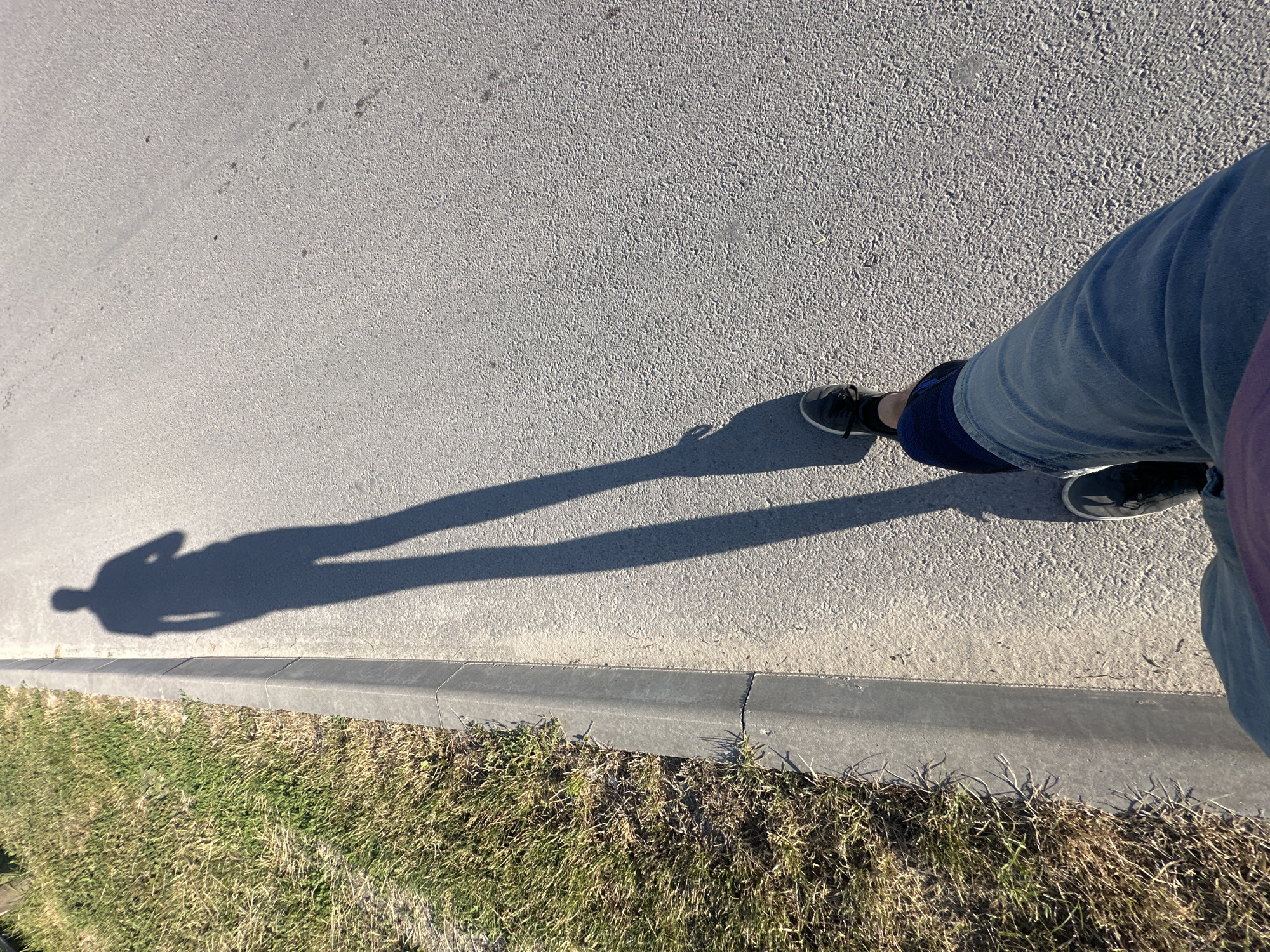 An individual's shadow is cast on a paved road, indicating a sunny day. The shadow is long and extends across the road, suggesting the sun is low in the sky. The individual's legs are visible in the foreground, wearing blue jeans and dark shoes. To the left of the road is a grassy area with a concrete curb. The scene appears to be outdoors, possibly in a park or along a street in the city of Makiv, Ukraine. The asphalt road surface is textured and shows some small debris. The grass is dry and somewhat sparse in places.