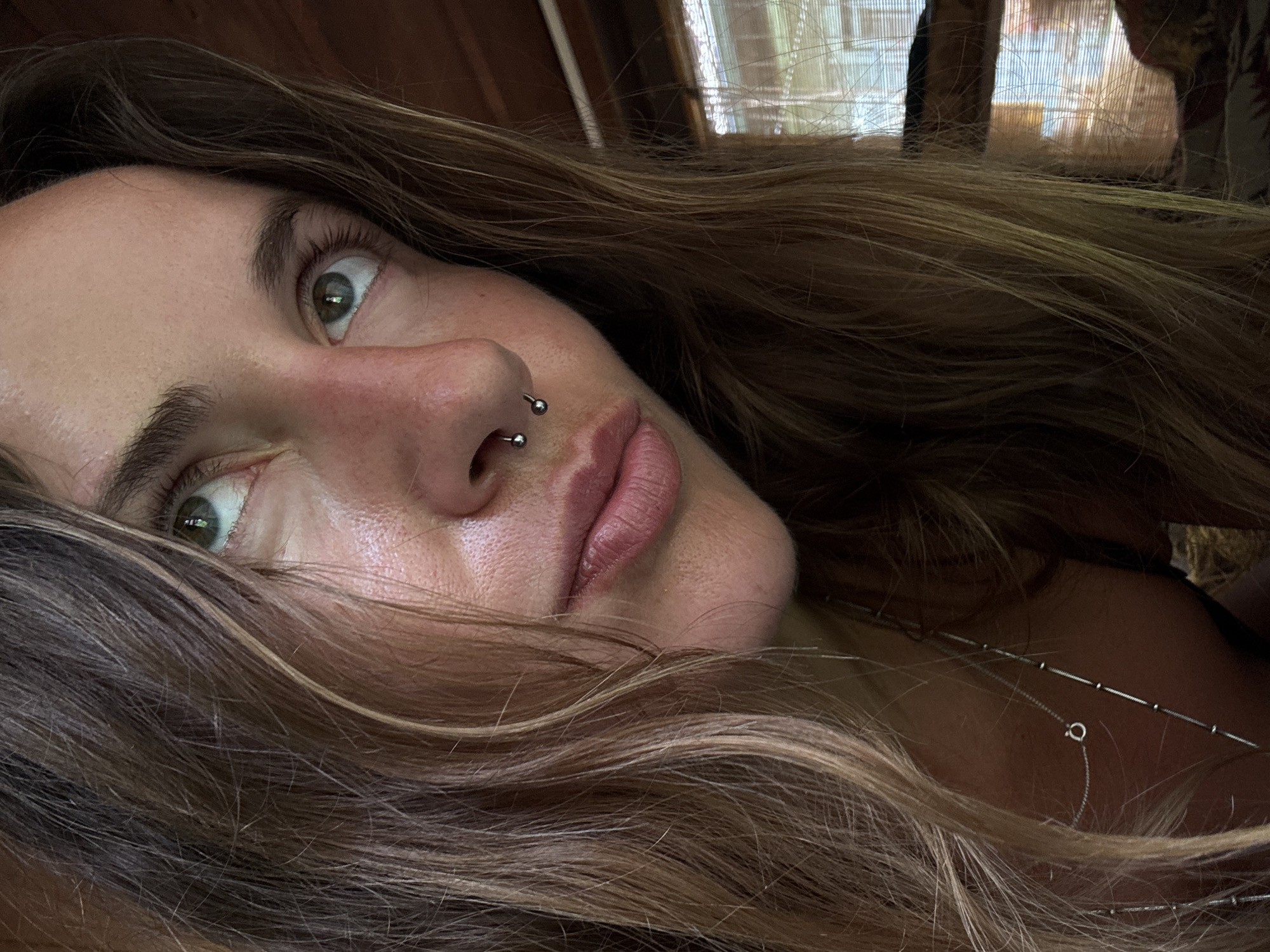 This is a close-up, angled selfie of a person with long, wavy, light brown hair. The person's face is partially obscured by their hair, with their left eye and nose prominently visible. They have green eyes and a septum piercing. Their lips are slightly parted, and their skin appears flushed. The background is out of focus, but appears to be an indoor setting with wooden elements and a window with blinds. The lighting suggests it could be daytime. The person's expression is difficult to determine due to the angle and focus, but their wide-open eye could suggest surprise or alertness. There is a thin, silver chain necklace visible around their neck.