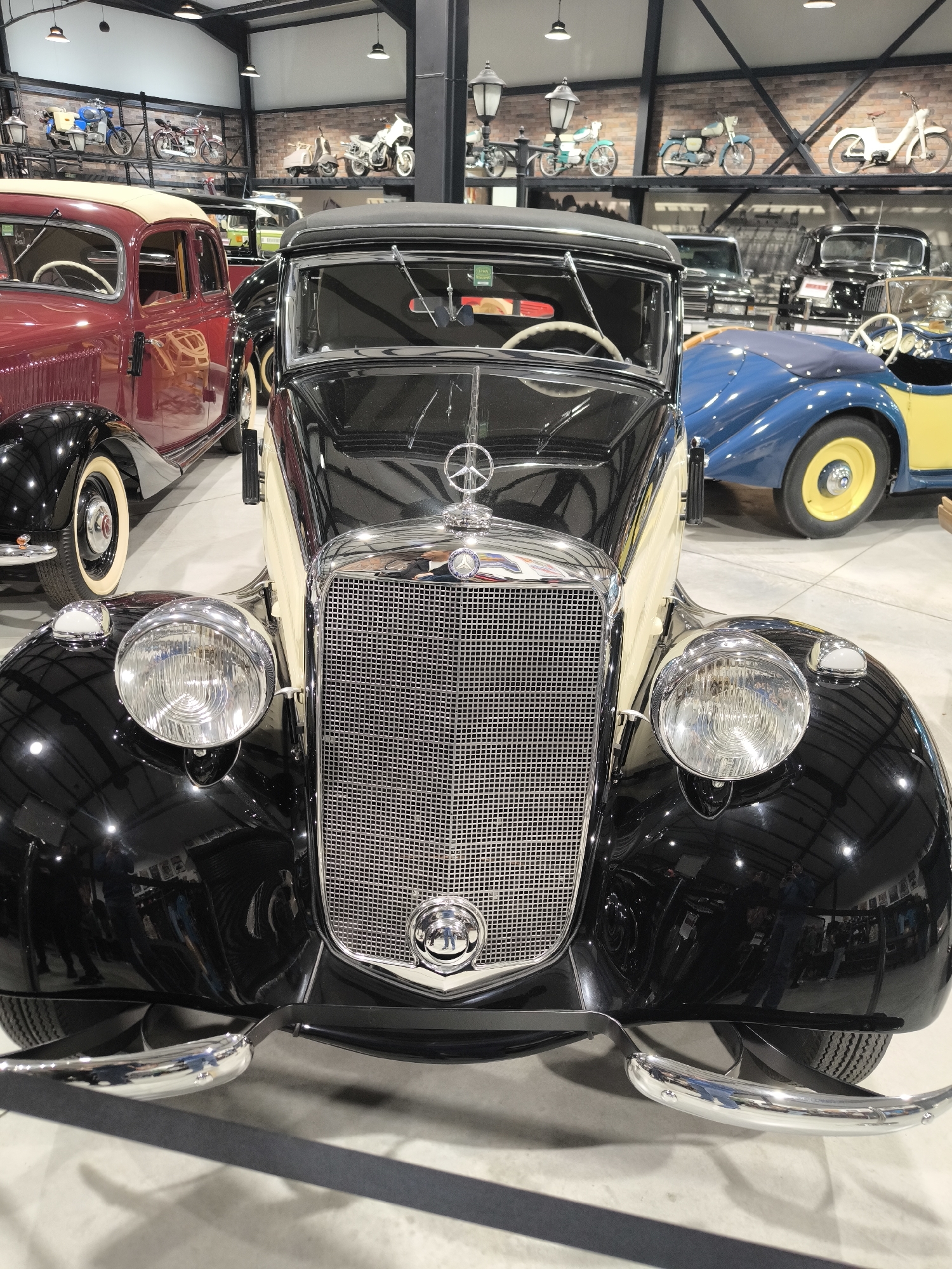 The image shows a black and cream-colored vintage Mercedes-Benz car in what appears to be a museum or showroom. The car has a prominent chrome grill with the Mercedes emblem. In the background, there are other classic cars and motorcycles displayed on shelves. The environment is well-lit and appears to be indoors. There's no definitive evidence to confirm the exact location in Kapatovo, Bulgaria, without additional context.