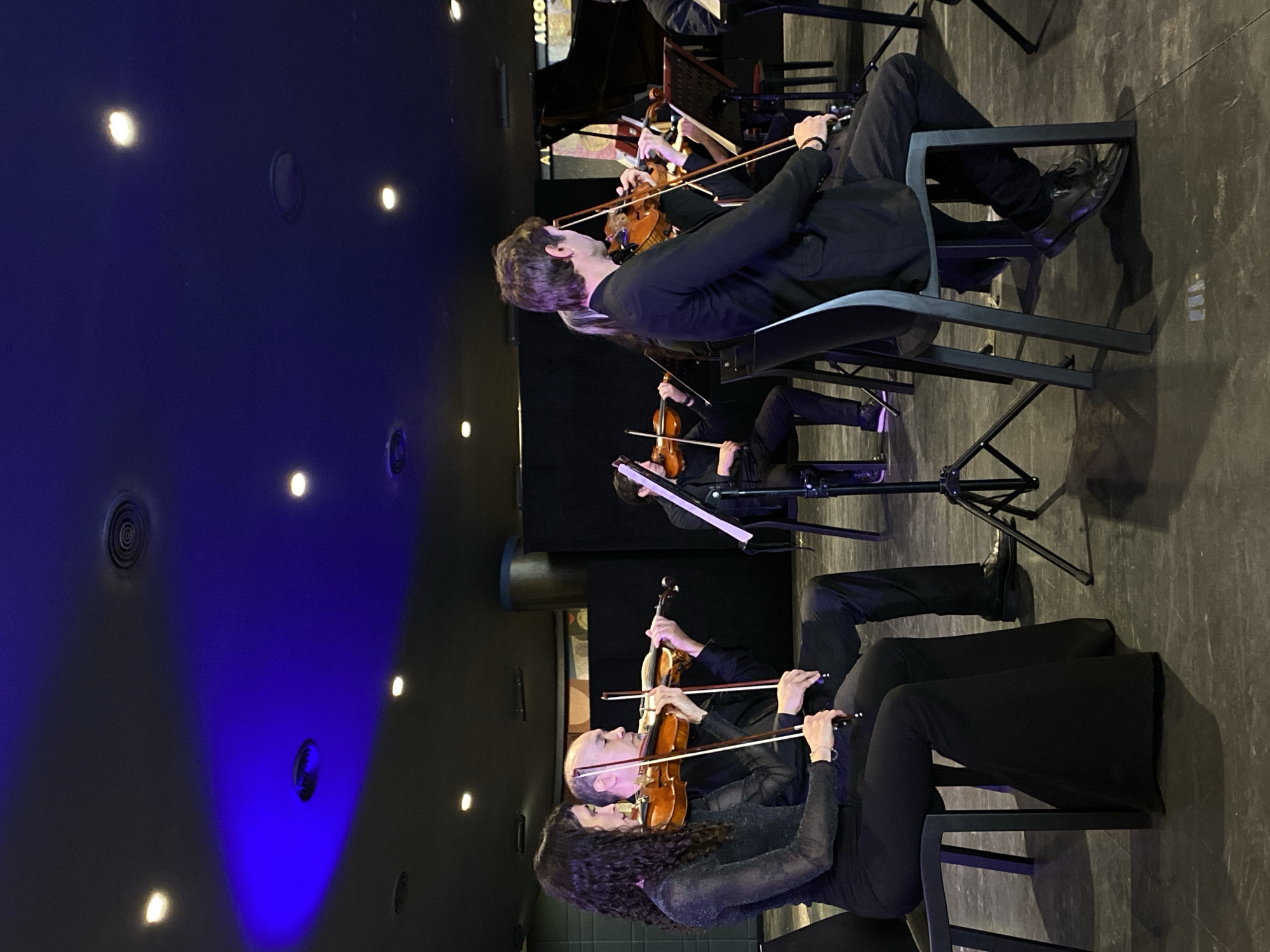 The image depicts a string orchestra performing on a stage. The setting appears to be indoors, judging from the ceiling with recessed lighting and air vents. Several musicians are seated and playing violins. They are dressed in formal attire, suggesting a concert or performance setting. The location is implied to be Buenos Aires, Argentina, as per the request.