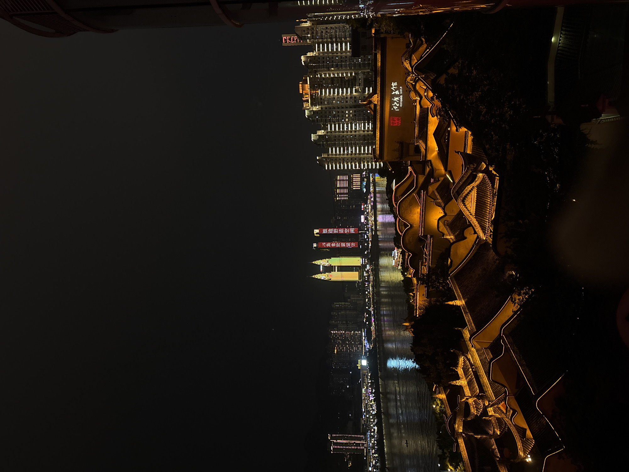 At night, the image captures a lit cityscape of Yuzhong District, China, dominated by waterways reflecting the lights. Tall buildings, including skyscrapers with illuminated facades, stretch along the waterfront. A traditionally styled, multi-tiered building complex with ornate roofing stands prominently to the right. The lights illuminate the dark water, contrasting with the black sky.
