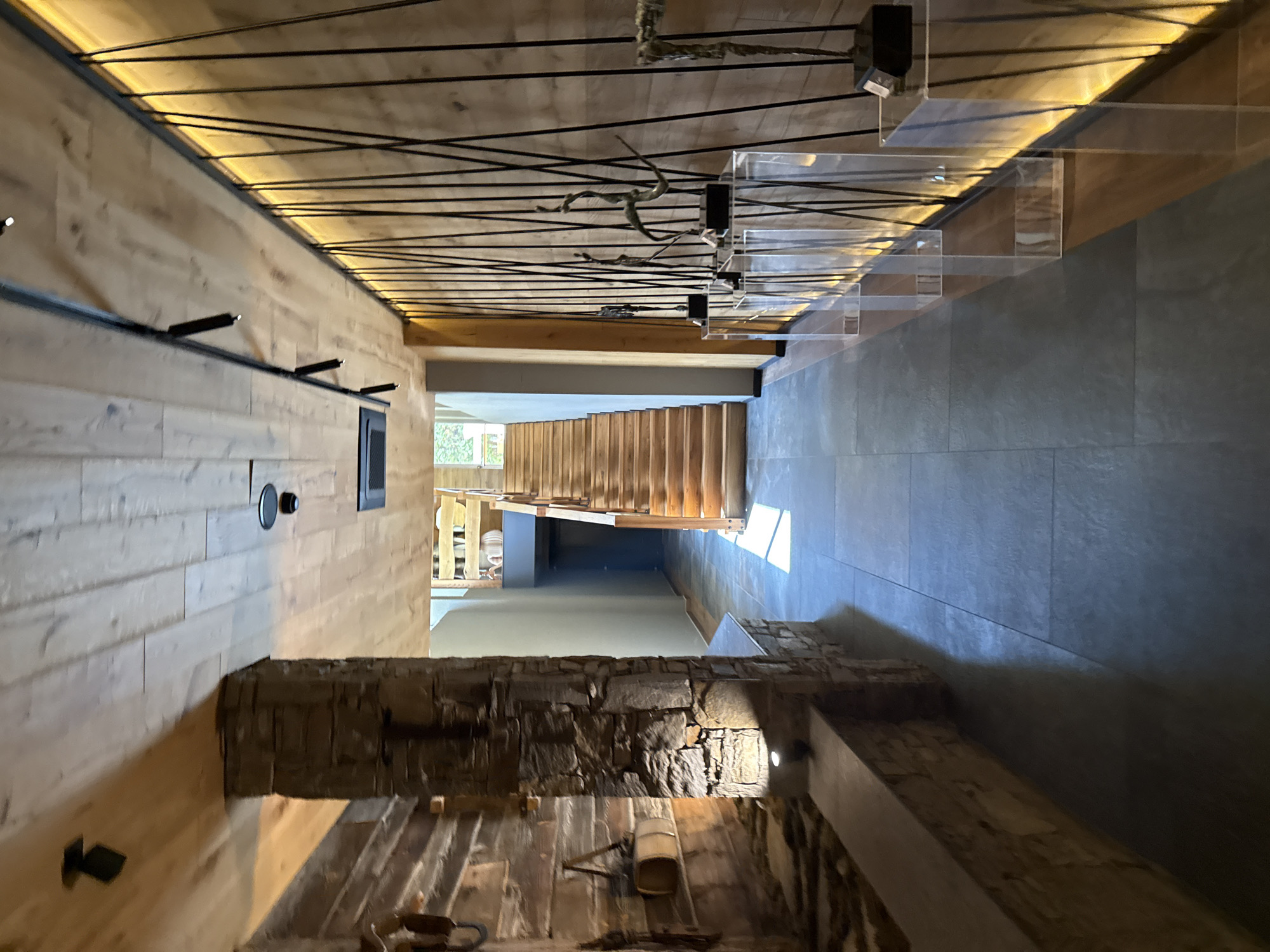 The image depicts the interior of a building in Velingrad, Bulgaria. The viewpoint is upward towards the ceiling, revealing a wooden slat ceiling with a black linear design. A hallway is visible with tiled floors and a staircase leading upwards. The stairs have transparent railings. The walls are a mix of wood paneling and exposed stone or brickwork. A window is present in the distance, allowing natural light to enter.
