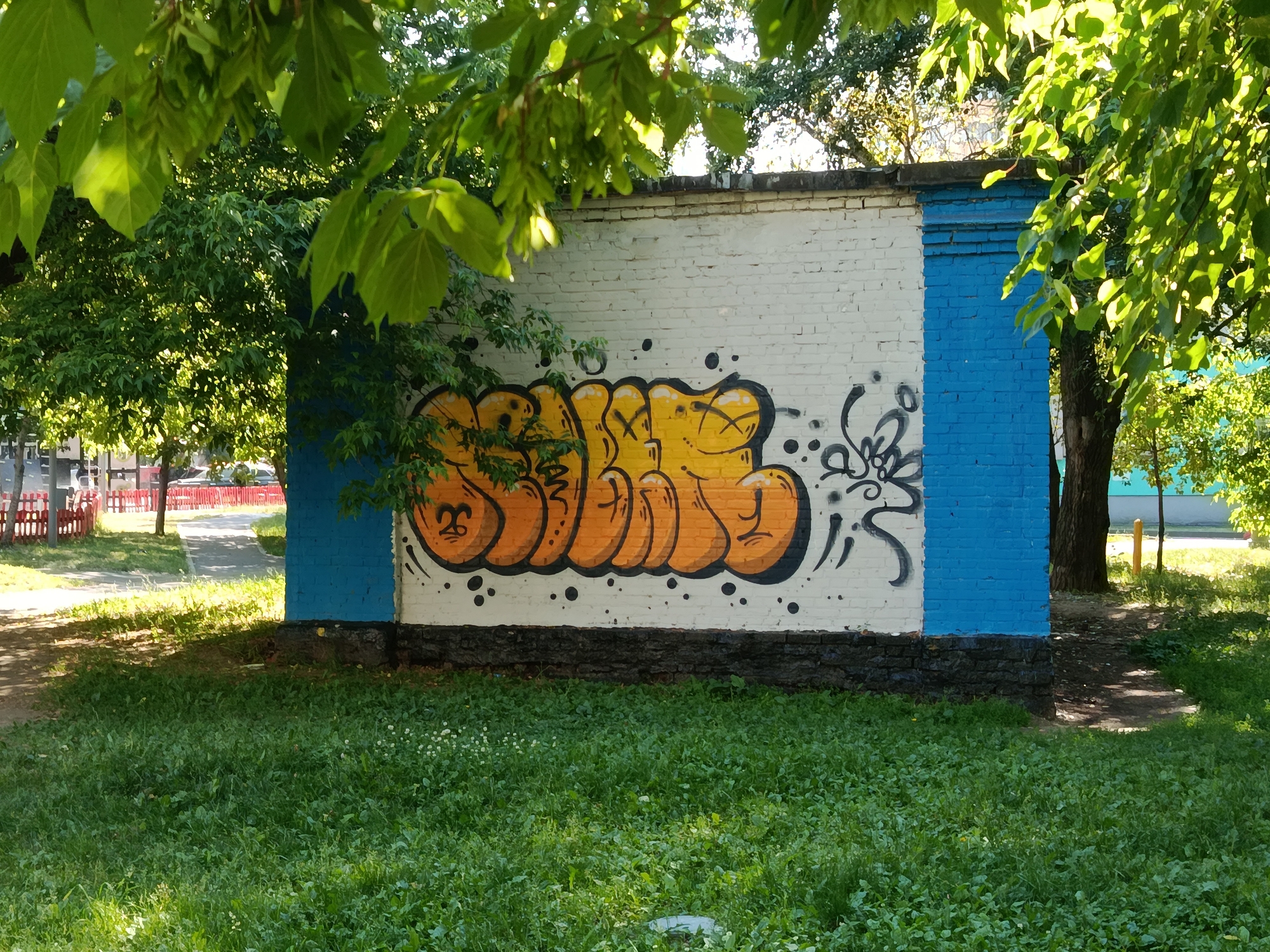 The image depicts a section of a brick wall in an urban park setting, likely in Moscow, Russia. The wall is painted white on the left half, with a large graffiti tag in yellow and orange dominating this section. The graffiti features bubble-like letters with black outlines and some details resembling eyes or crosses. To the right of the white section, the wall is painted a vibrant blue. The lower portion of the wall has a dark, textured base, possibly stone or weathered brick.
The wall is partially obscured by the lush green leaves and branches of trees in the foreground, casting dappled sunlight onto the scene. The ground in front of the wall is covered in thick green grass, with a few small white flowers scattered throughout. In the background, a pathway is visible, lined with more greenery and a red fence. Beyond the fence, there are glimpses of buildings and parked cars, suggesting the park is situated in a residential or mixed-use area. The overall impression is a sunny, outdoor environment.