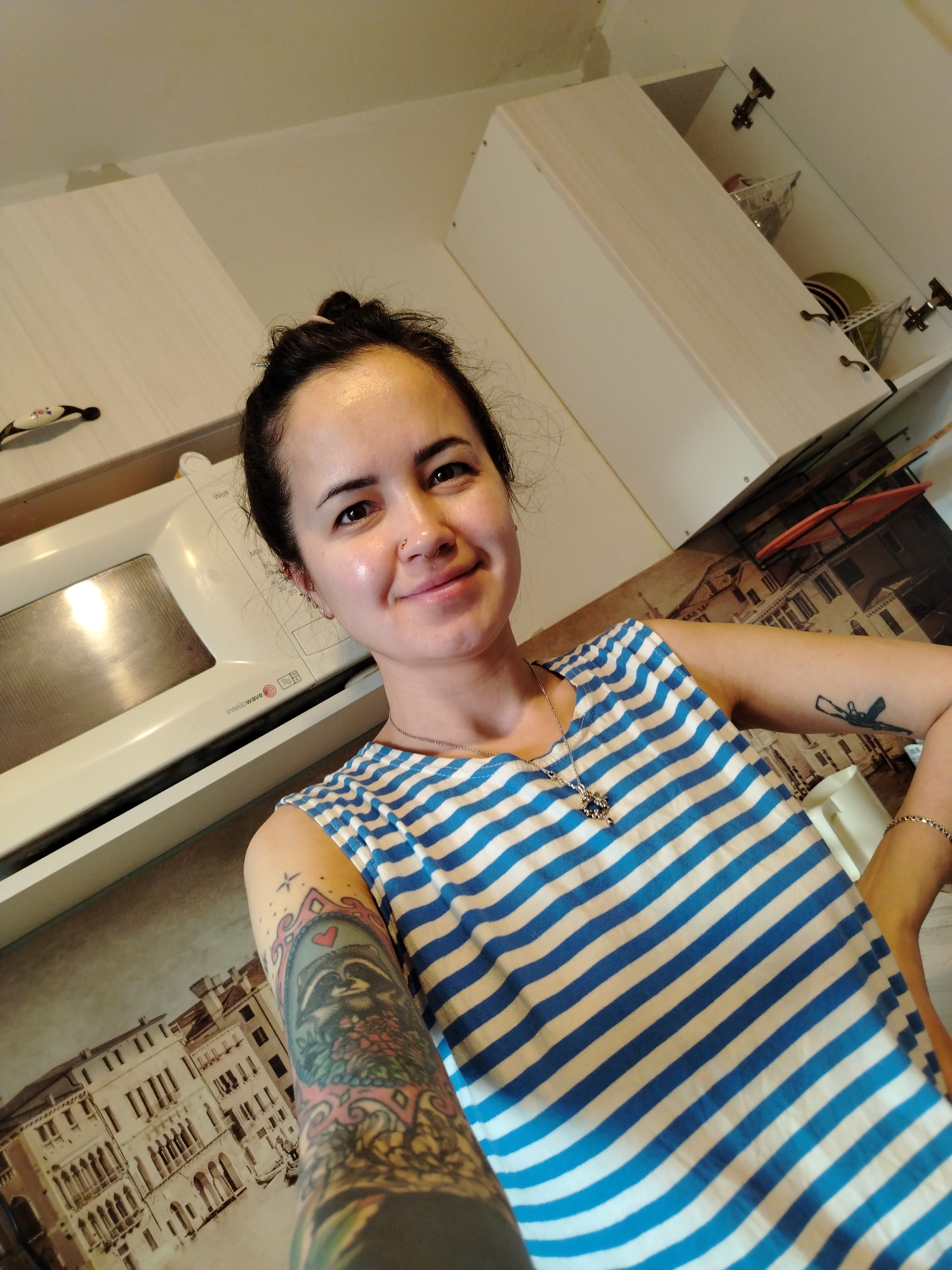 The image captures a smiling woman posing for a selfie indoors, likely in a kitchen in Noviy, Russia. She has dark hair styled in a bun, a nose piercing, and multiple earrings. She wears a blue and white striped sleeveless top, a silver necklace with a decorative pendant, and a thin silver bracelet on her right wrist. Her left arm is extensively tattooed with colorful designs, including a raccoon depicted within an ornate frame. A smaller, dark, abstract tattoo is visible on her right upper arm.
The background features white upper kitchen cabinets, some partially open revealing dishes, including stacked yellow/green plates and clear plastic containers. To her left, a white microwave oven is visible, displaying the text "intellowave," "Watts," and "Min," along with other control panel markings. The wall or backsplash behind her features a decorative print depicting traditional buildings, reminiscent of European architecture. The lighting appears bright and natural, indicating daytime. The woman's expression is pleasant and relaxed.