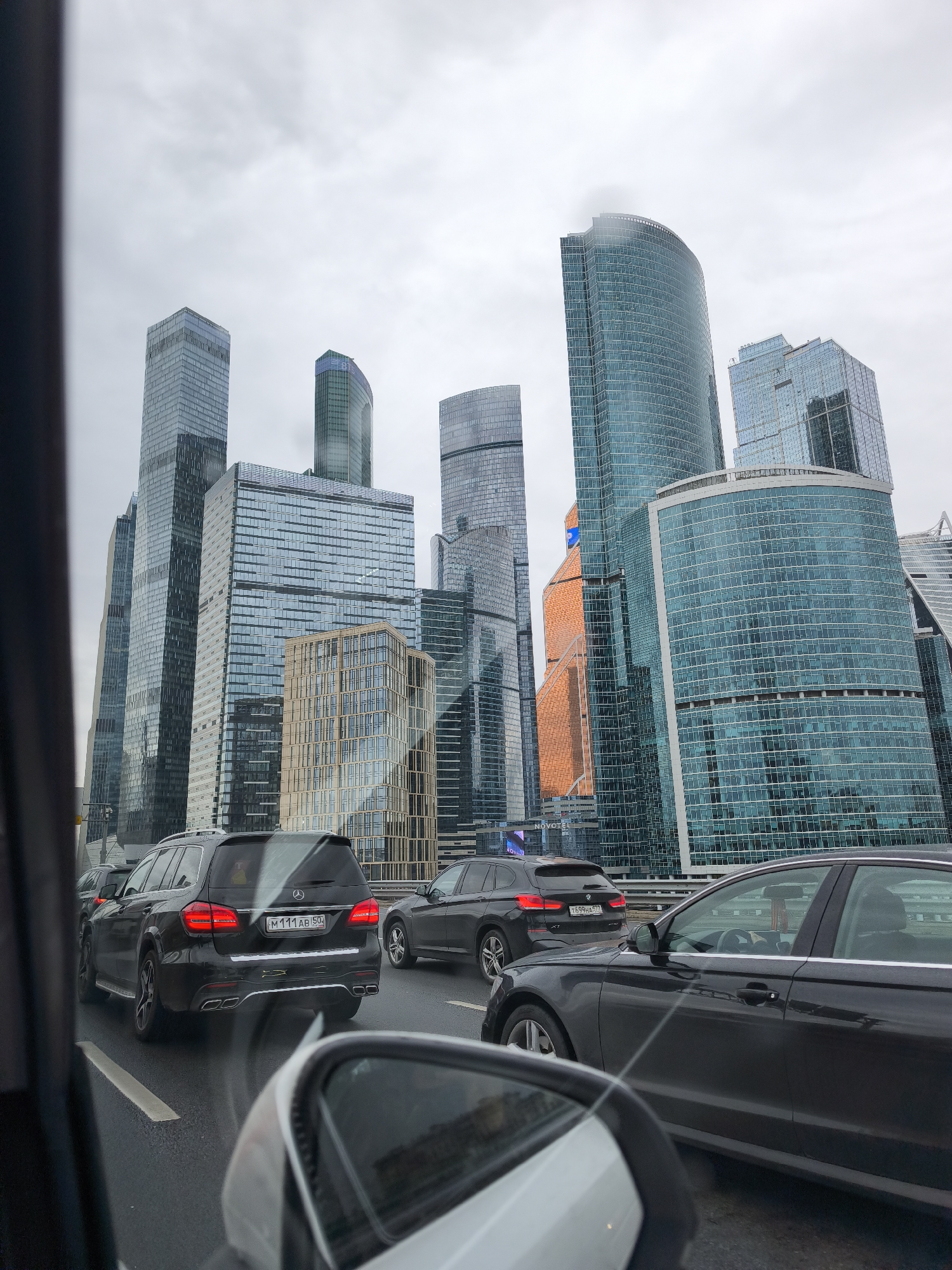 The image, taken from inside a vehicle on a bridge, depicts a street scene in Moscow, Russia, featuring the Moscow International Business Center ("Moscow City") skyline. Several modern skyscrapers of varying heights and designs are visible, including glass-covered towers. A gray, overcast sky is overhead. Three dark-colored cars are on the bridge in traffic; the first is a Mercedes-Benz GL-Class SUV, license plate "M111AB 50," followed by a BMW X Series. The third car is an Audi. The side mirror of the vehicle from which the photo was taken is visible in the lower-left corner of the frame. The road appears to be wet, suggesting recent or ongoing rain.